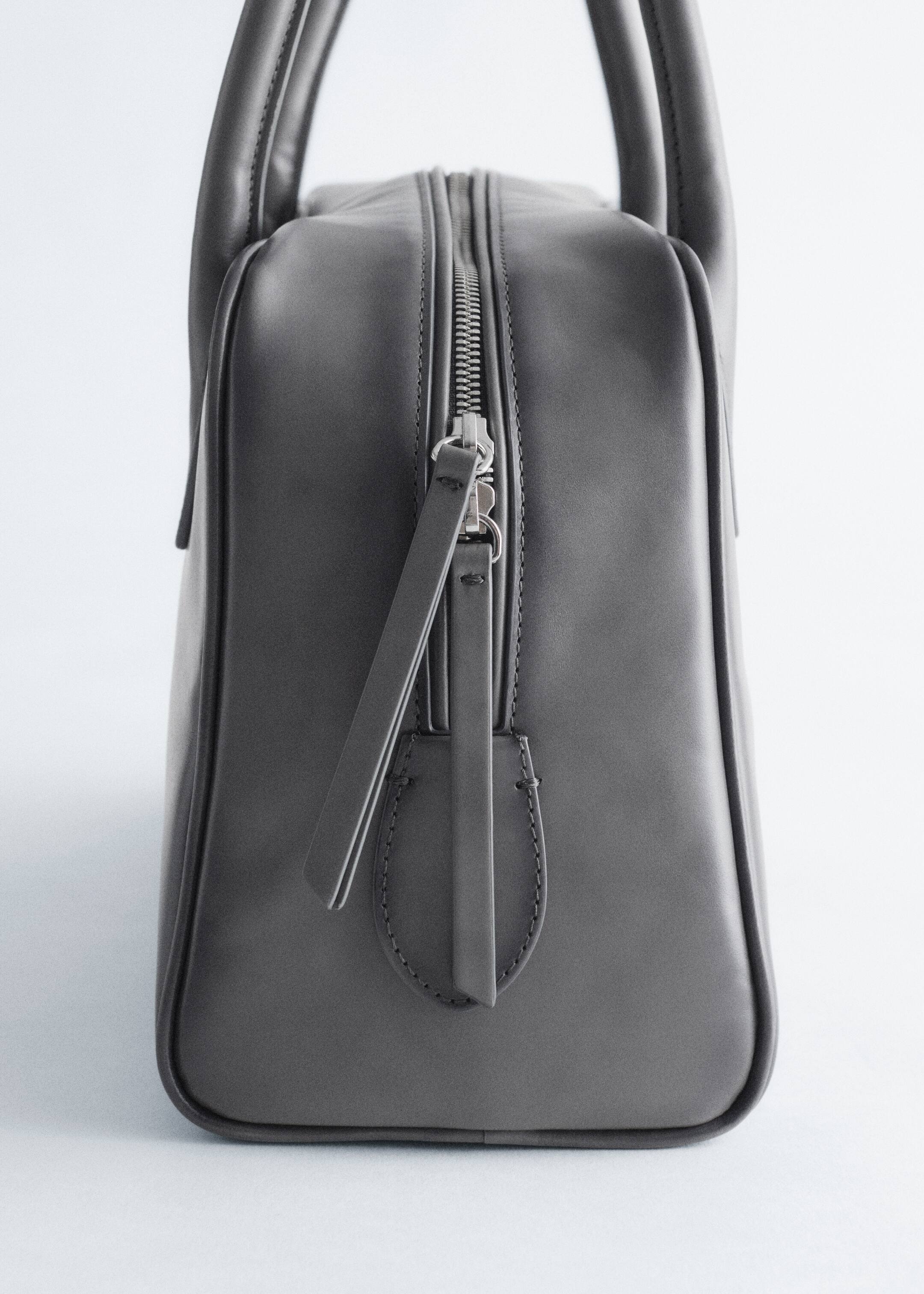 Grösseres Bild ansehen: Eine strukturierte dunkelgraue Ledertasche ist nach links ausgerichtet und verfügt über einen durchgehenden oberen Reißverschluss mit einem silberfarbenen Zipper und einem passenden Lederband. Zwei abgerundete obere Griffe ragen vertikal aus der Tasche heraus.