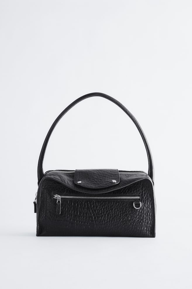 Un sac à bandoulière en cuir texturé noir est présenté de face, avec une ligne supérieure incurvée et un rabat central fermé par deux boutons-pression argentés. Une seule bandoulière arquée sâélève vers le haut, et une poche horizontale zippée avec des finitions argentées détaille lâavant. - 1
