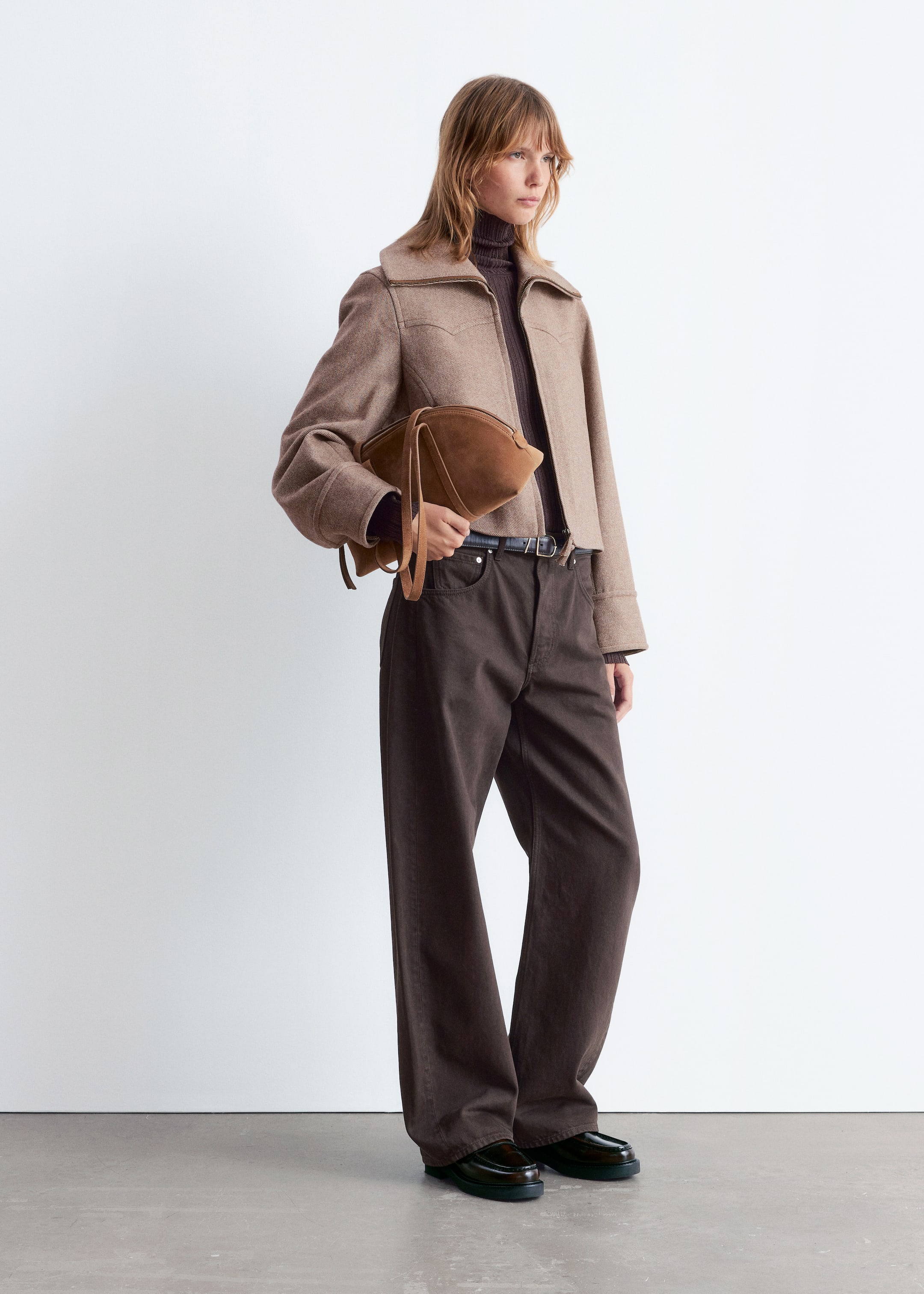 Größeres Bild ansehen: Ein Model präsentiert ein Outfit in neutralen Tönen und blickt leicht nach rechts. Eine hellbraune, kurze Jacke mit spitzem Kragen und westlichen Schulterjochen wird über einem dunkelbraunen, gerippten Rollkragenpullover getragen, kombiniert mit dunkelbraunen, weit geschnittenen Hosen, einem dunklen Gürtel und dunklen, klobigen Loafern. Eine braune, unstrukturierte Clutch wird gehalten.