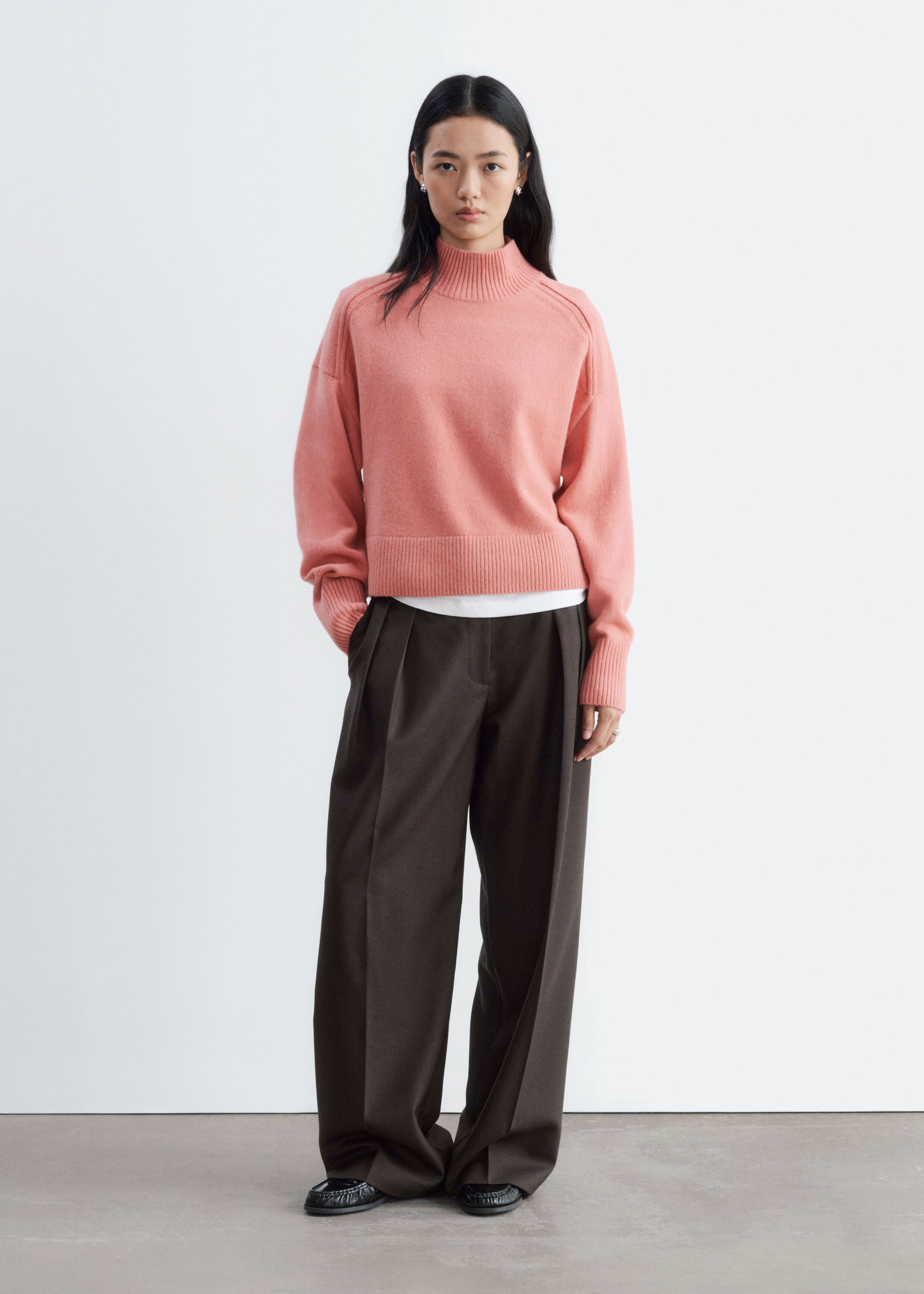 The model is wearing a pink mock neck sweater featuring a ribbed collar, cuffs, and hem with forward-rolling shoulder seams. The knitwear is layered over a white T-shirt and styled with dark wide-leg trousers.