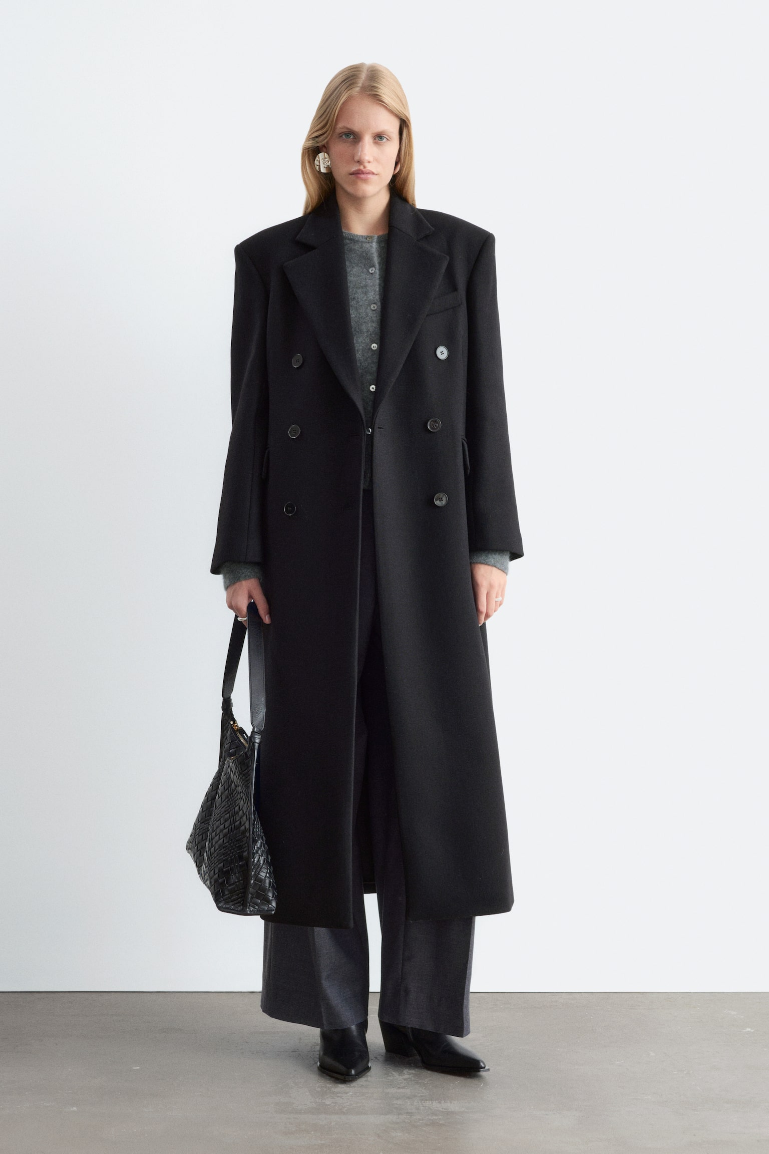 Ein schwarzer, zweireihiger Mantel aus Wollmischung wird VORNE präsentiert, mit einer langen, geraden Silhouette und gepolsterten Schultern. Er ist mit Kerbrevers, einer einzelnen Leistentasche auf der Brust, zwei Leistentaschen an den Seiten und geknöpften Manschetten gestaltet. - 1
