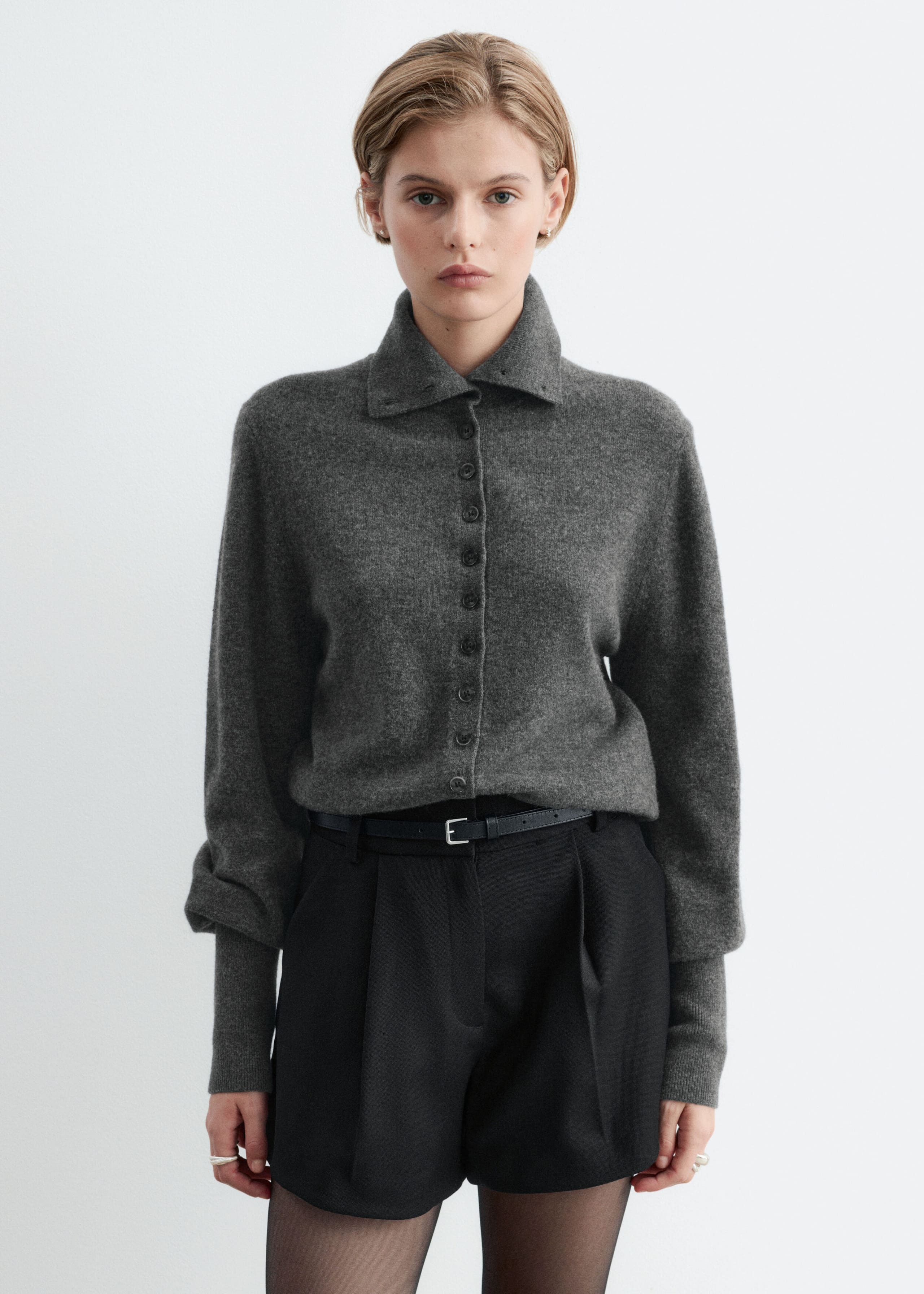The model is wearing high-waisted black shorts featuring front pleats and a soft A-line silhouette. The shorts are styled with a slim black leather belt, sheer black tights, and a tucked-in grey button-through knit shirt.