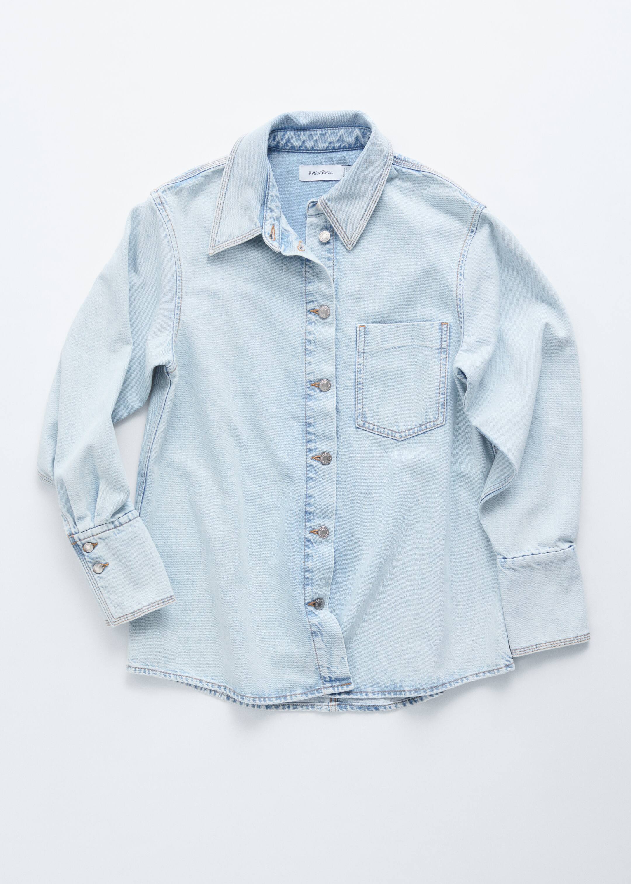 View larger image: A light blue denim shirt is presented front-facing, featuring a pointed collar, a full snap-button placket, and light contrasting stitching. It is showcasing a single patch pocket on the left chest and long sleeves with a double-button cuff on the left and a folded cuff on the right.