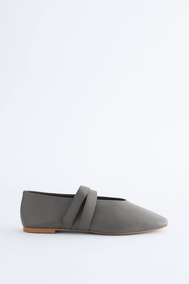 A grey flat shoe is displayed facing LEFT, featuring a square toe and a light brown sole. A strap is crossing over the instep, fastening with a hook-and-loop closure. - 1