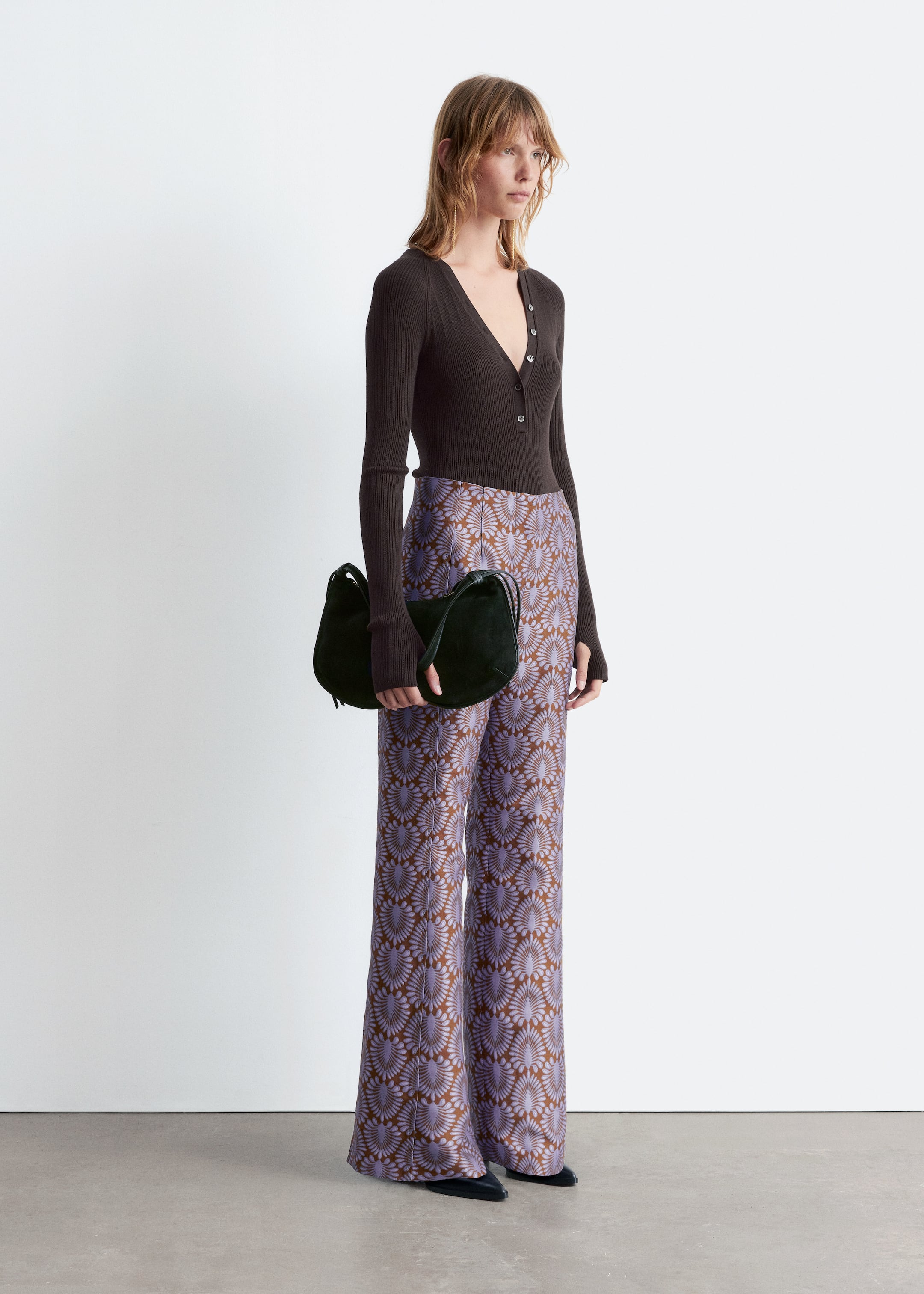 Visualizza immagine più grande: L'outfit presenta un top a V in maglia a coste marrone scuro con abbottonatura, abbinato a pantaloni a vita alta e gamba larga. I pantaloni mostrano un motivo a ventaglio ripetuto in viola chiaro e marrone, con una borsa a forma di mezzaluna verde scuro tenuta in mano e scarpe piatte a punta nere che completano l'insieme.
