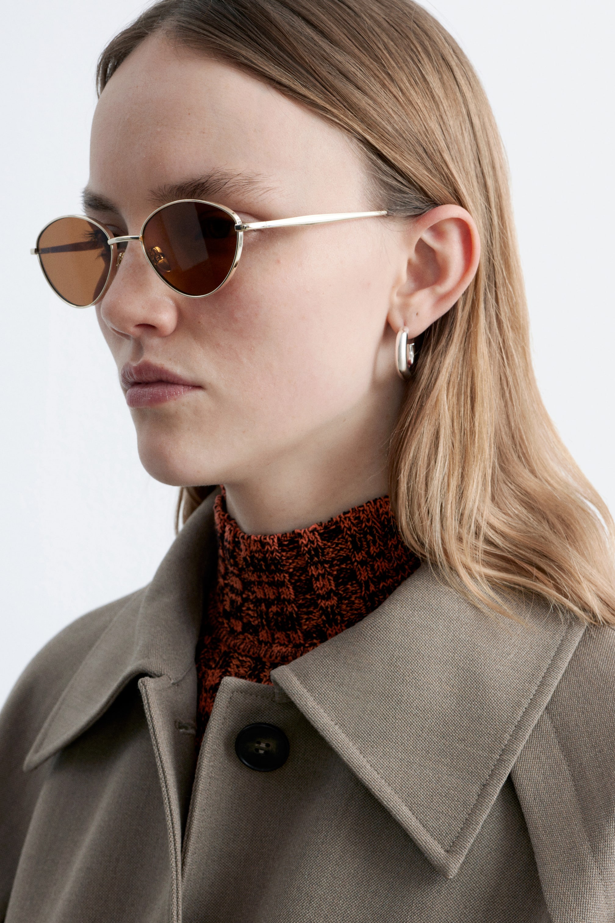 Grotere afbeelding bekijken: Goudkleurige ovale zonnebril met bruin getinte glazen wordt gedragen, iets naar links gekanteld, naast een zilverkleurige dikke hoepeloorbel. Een oranje en zwart gestructureerde gebreide top met een hoge hals wordt gelaagd onder een kaki jas, die voorzien is van een brede gestructureerde kraag en een donkere knoop.