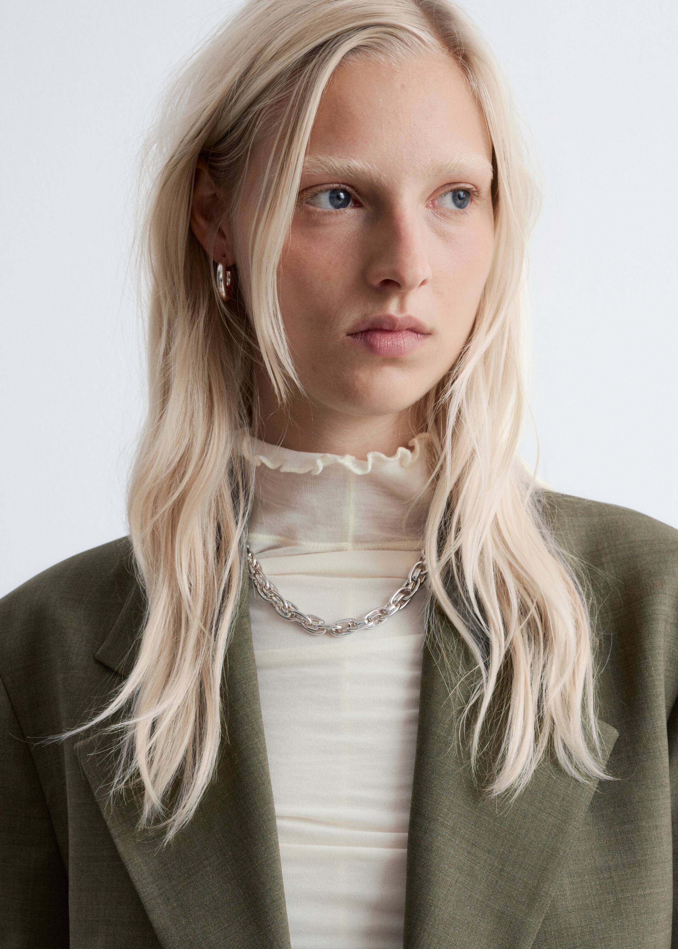 Visualizza immagine più grande: Un blazer verde oliva, caratterizzato da revers dentellati e una sottile texture intrecciata, è indossato aperto sopra un top color crema trasparente con collo alto e colletto arricciato. Una catena grossa color argento è posata sul top e orecchini a cerchio color argento adornano le orecchie della modella, tutti presentati con un leggero angolo.