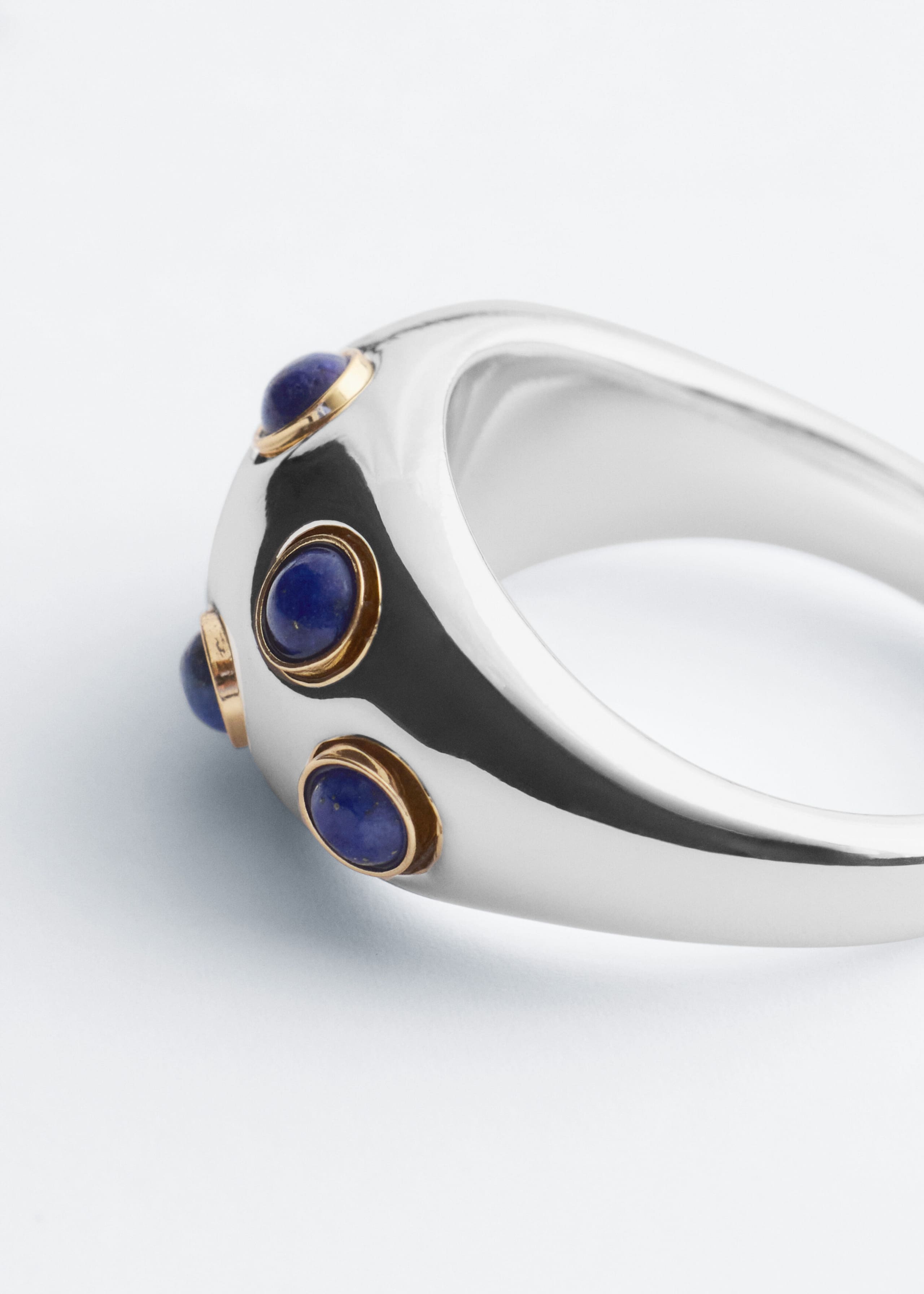 Ein polierter, silberfarbener, skulpturaler Ring liegt schräg und präsentiert sein breites, geschwungenes Band. Auf der Außenseite sind mehrere runde blaue Cabochon-Steine zu sehen, die in erhabenen, goldfarbenen Fassungen gefasst sind.