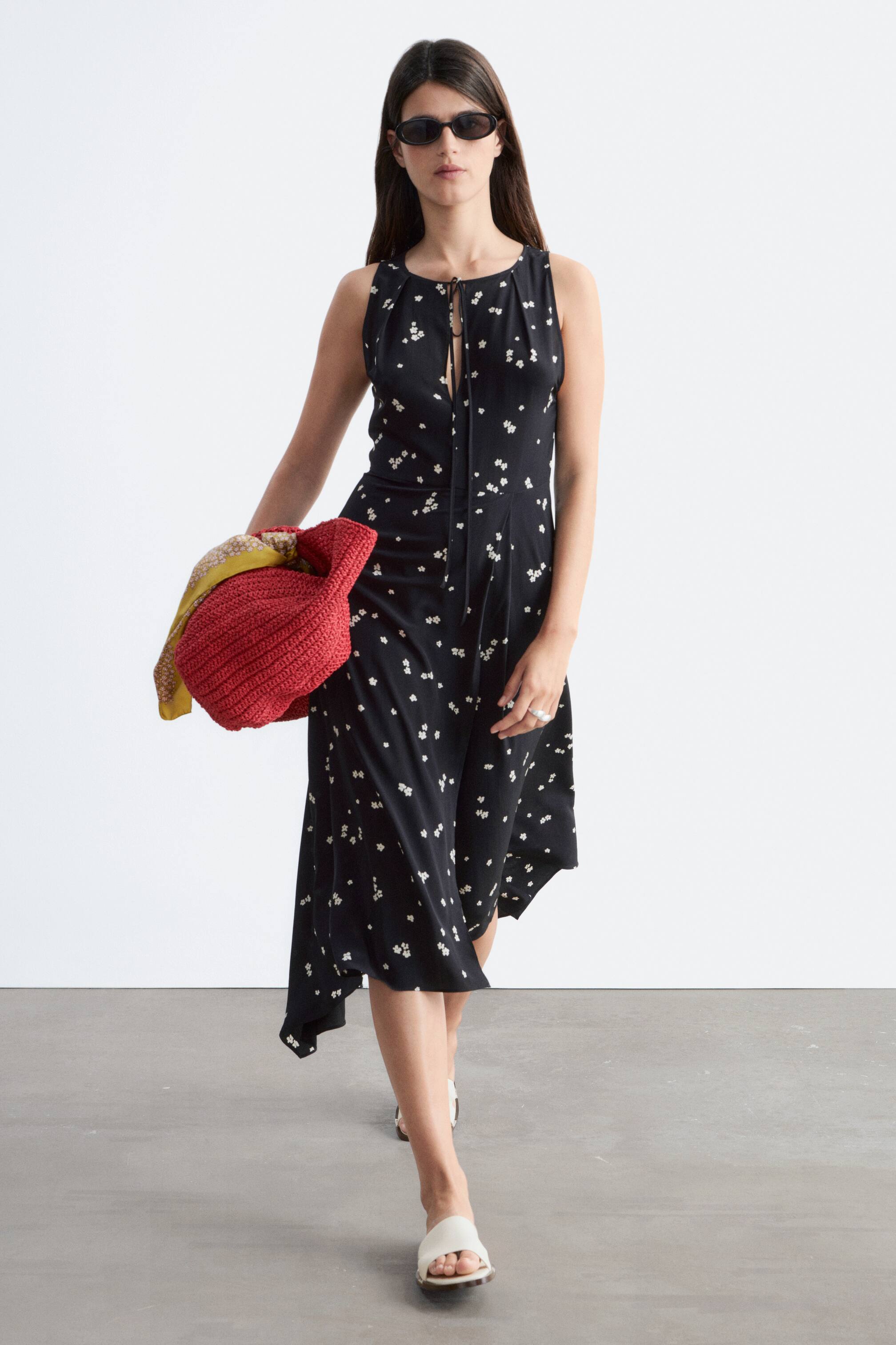 הצגת תמונה גדולה יותר: שמלה שחורה ללא שרוולים עם הדפס פרחים לבן מציגה מחשוף קי-הול עם קשרים, שולי מידי אסימטריים וסילואטה זורמת.