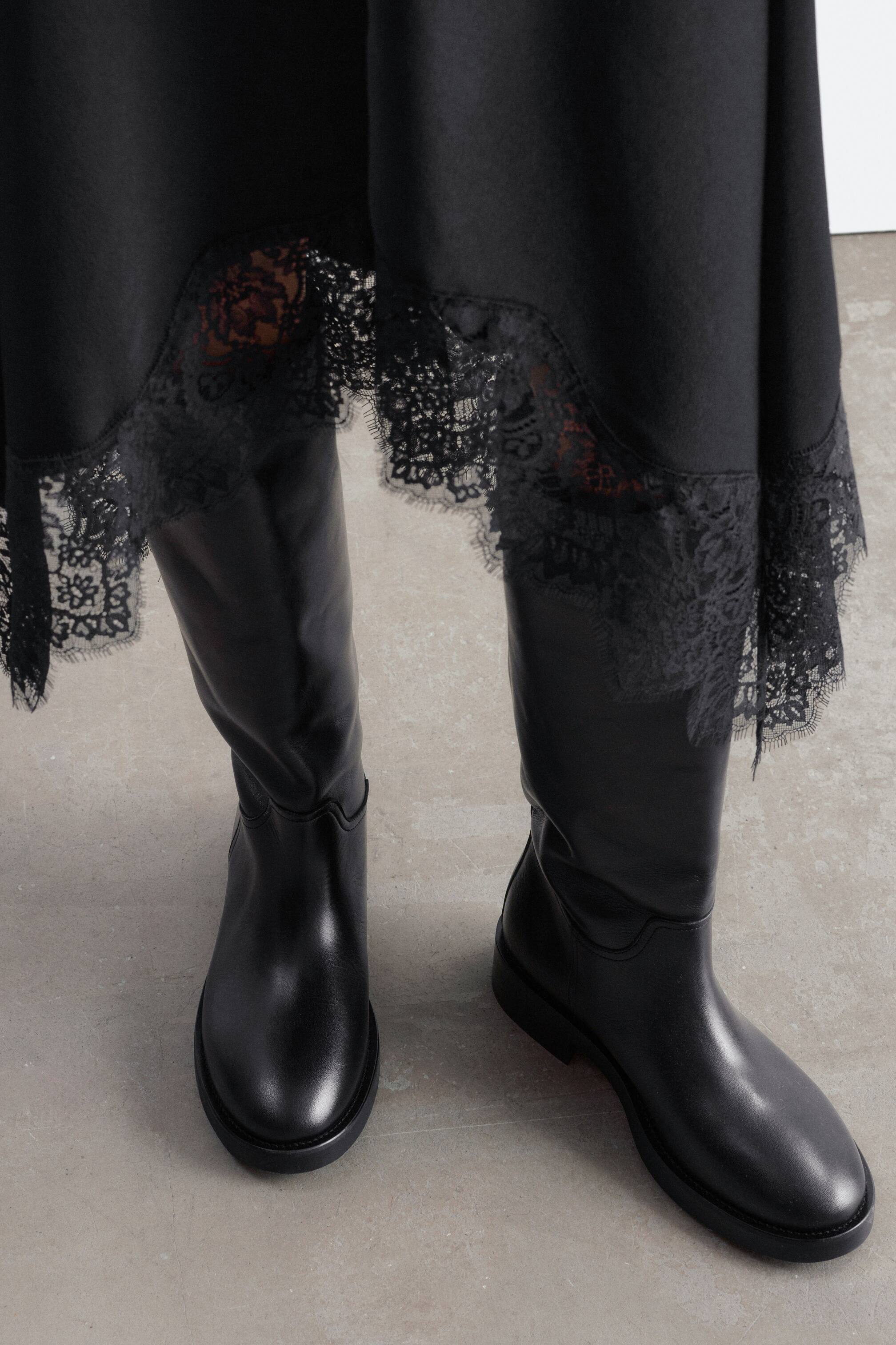 Agrandir l'image: Des bottes en cuir noir montant jusqu’au genou, dotées d’un bout arrondi et d’un petit talon bloc, sont portées par le mannequin. Les bottes présentent une finition lisse et légèrement brillante et sont associées à une jupe noire avec un ourlet bordé de dentelle.