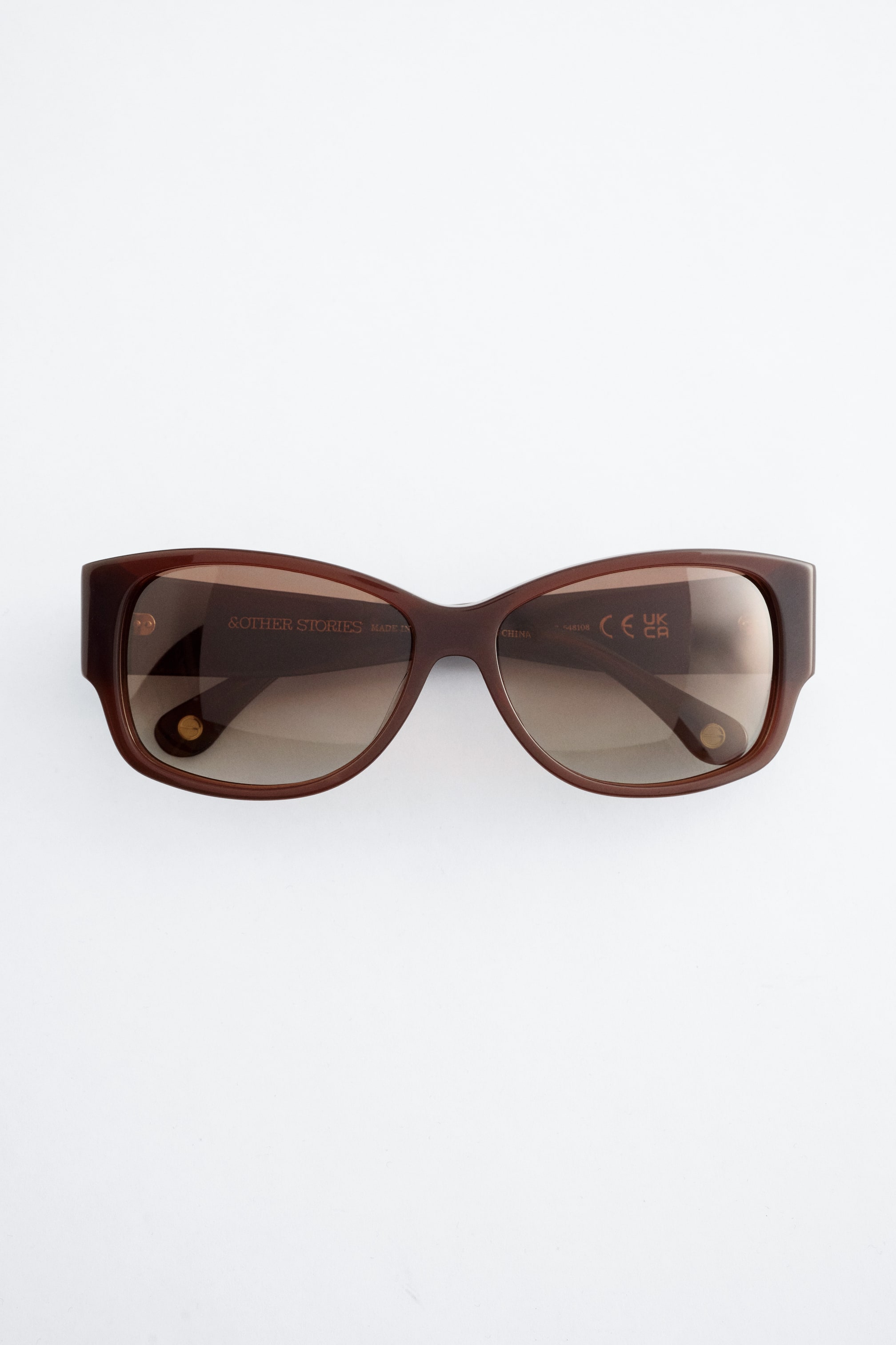 Ver imagem maior: Óculos de sol em acetato castanho estão virados para a frente, apresentando uma armação oval com bordas arredondadas, hastes largas e lentes com gradiente tonal castanho sombreado.