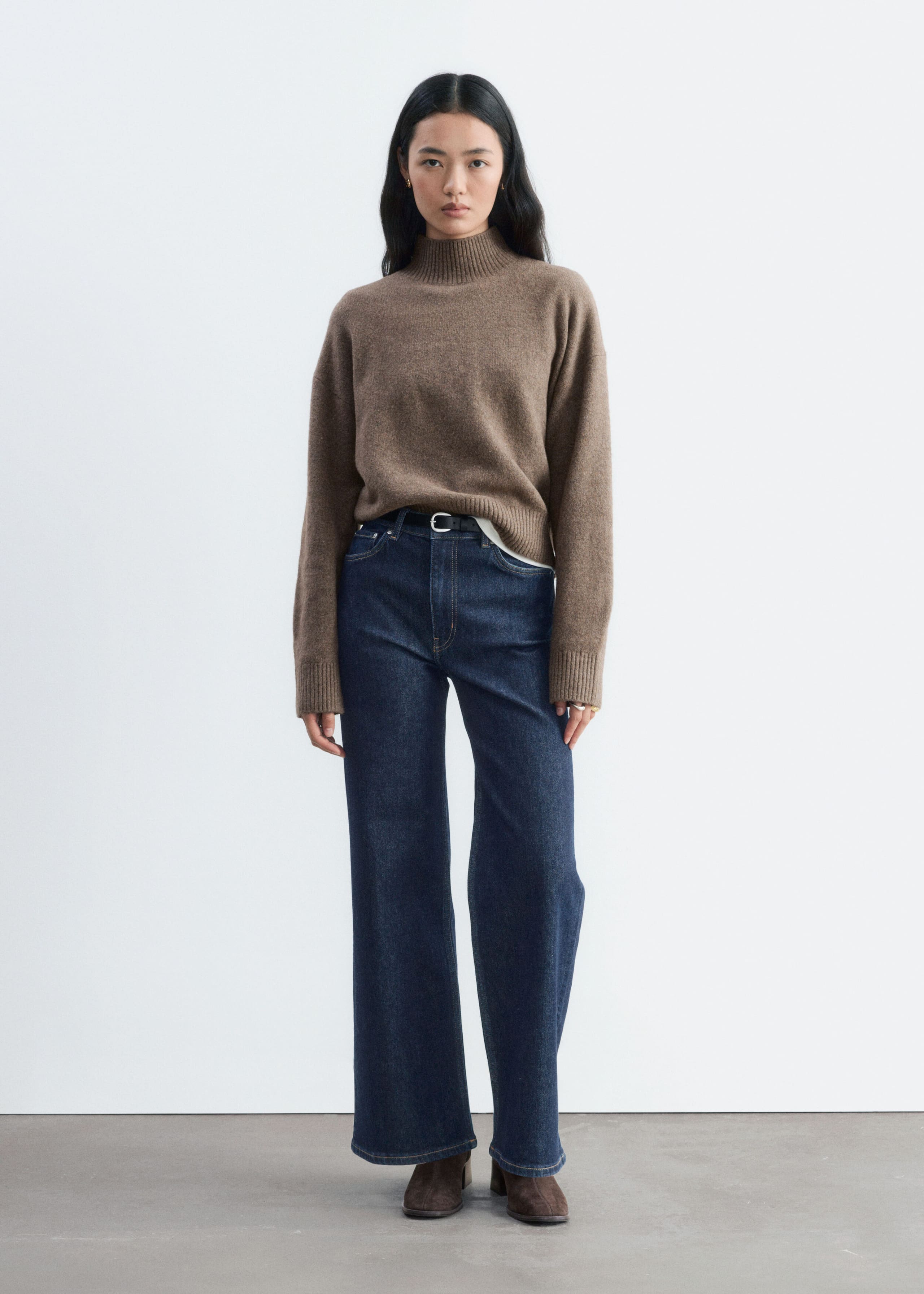 Een bruin gebreid trui met een geribbelde col, verlaagde schoudernaden en een losse pasvorm wordt door het model gedragen. De trui is in de taille ingestopt in donkerblauwe wide-leg jeans.