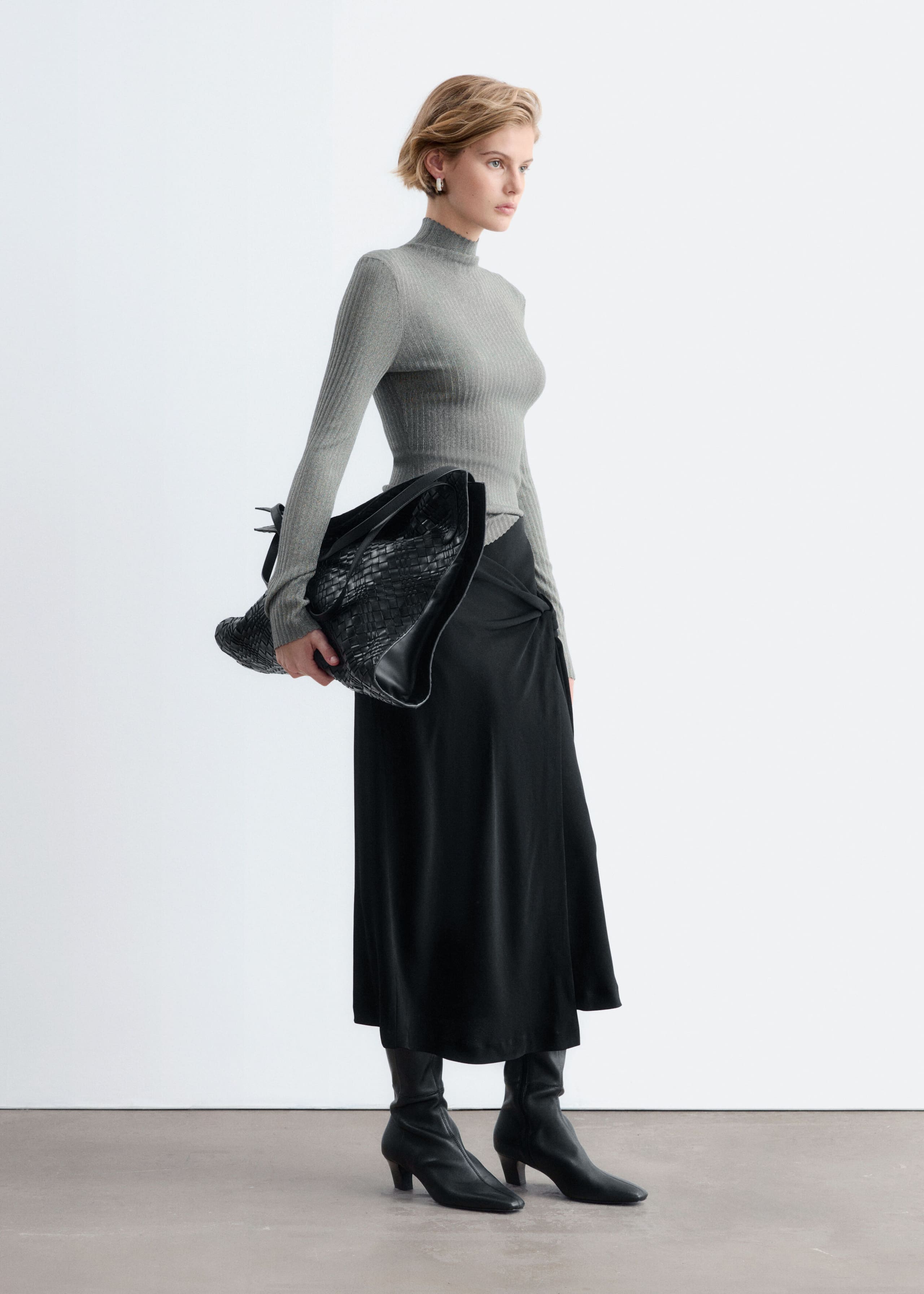 A gray, long-sleeved rib-knit top with a mock neck and a subtle metallic sheen is worn by the model, who is standing in profile. The top is styled with a black midi skirt, black leather boots, and a large woven black handbag carried under the arm.