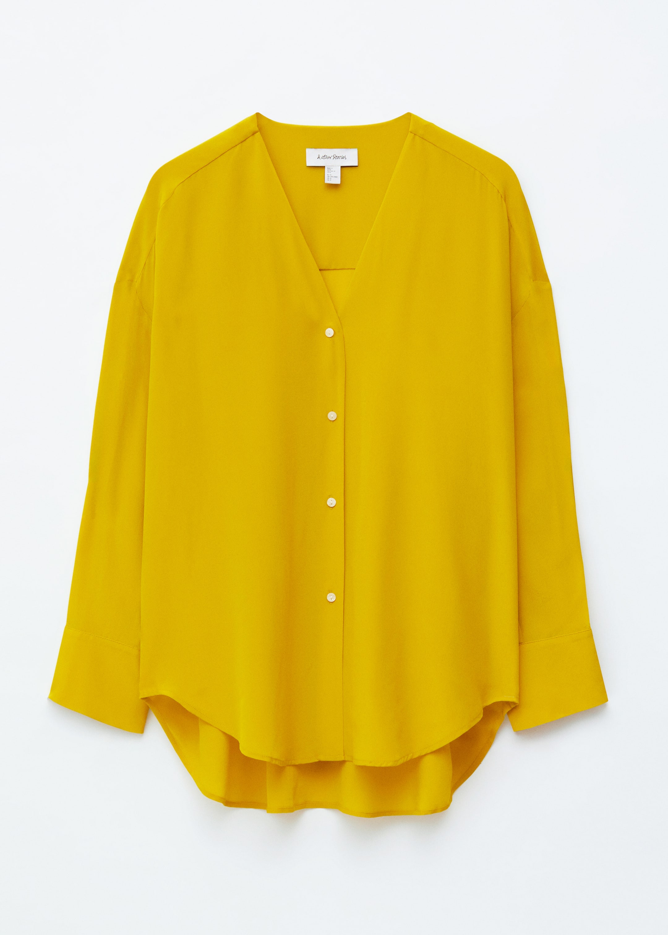 Ver imagen más grande: Una blusa amarilla presenta un escote en V con un panel subyacente, y su tapeta frontal está asegurada por cinco botones redondos de color claro. Las mangas largas y relajadas terminan en puños anchos, y un dobladillo curvo y asimétrico completa el diseño.