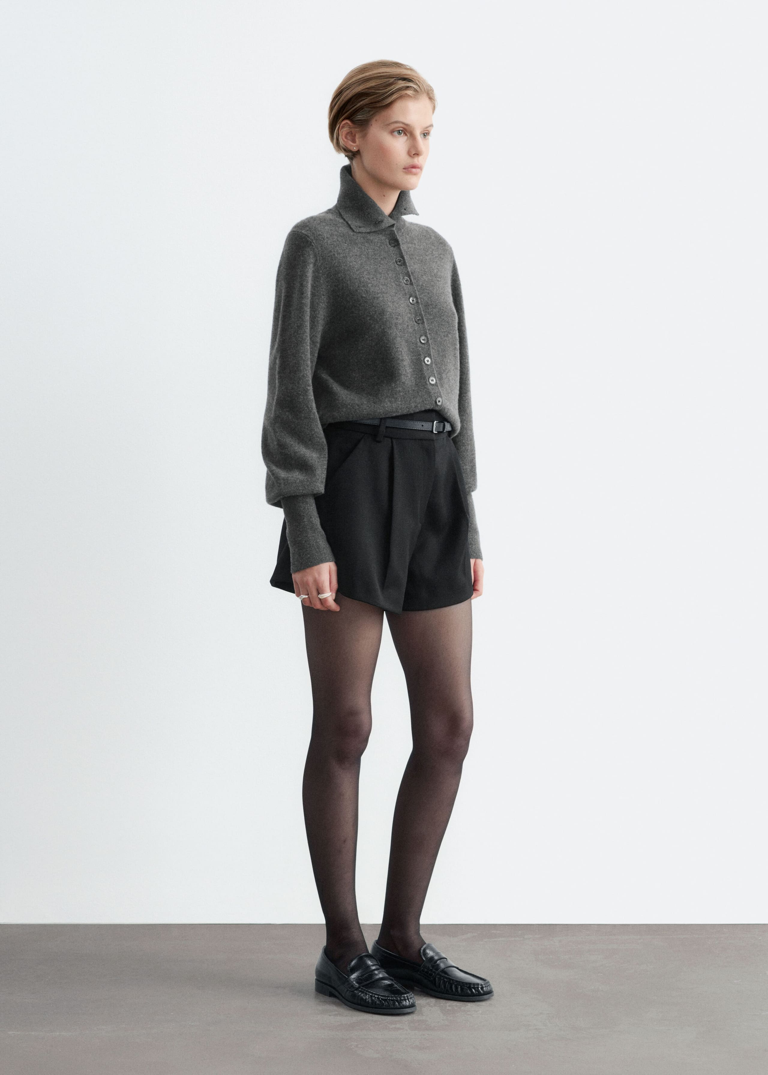 Black high-waisted shorts with front pleats and a subtle A-line silhouette are worn by the model. The shorts are styled with a thin black belt at the waist, a tucked-in grey knit button-up top, sheer black tights, and black loafers.