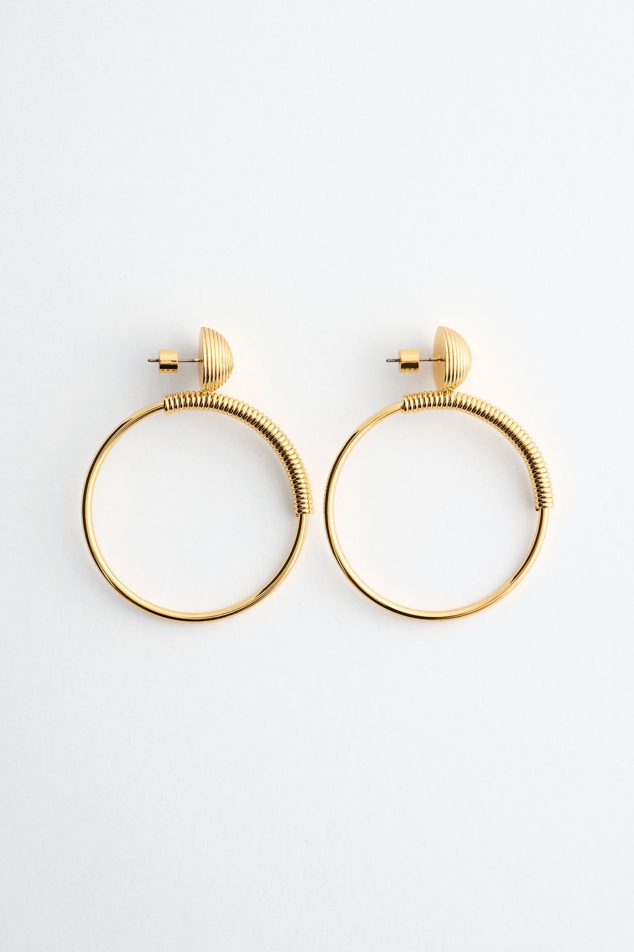View larger image: The Polished Brass Coil-Detail Hoops are shown from the front, featuring large circular hoops suspended from ribbed, semi-spherical stud backings. A fine coiled-wire detail wraps around the upper outer curve of each polished gold-toned hoop.