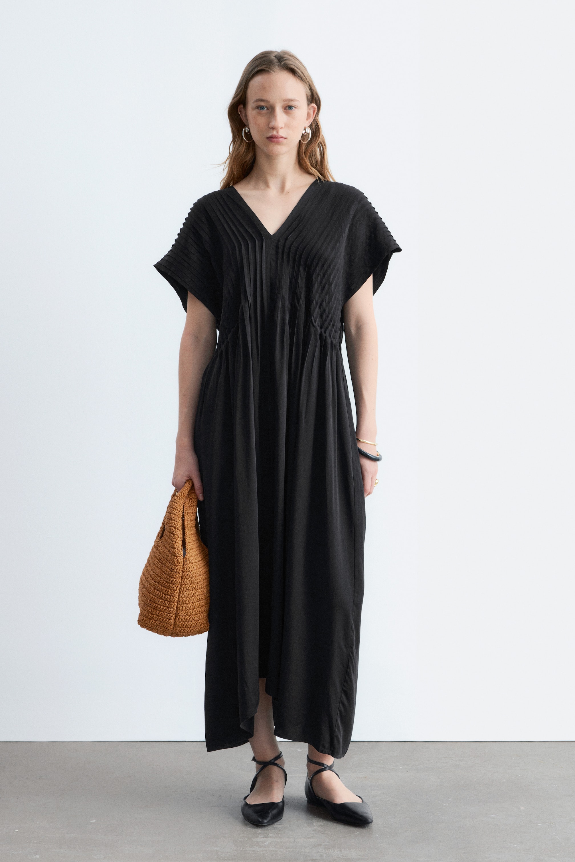 Se større billede: En sort midikjole med V-udskæring, korte brede ærmer og en forside med lodrette pintucks. En rynket talje flyder ud i et løst skørt med asymmetrisk kant.