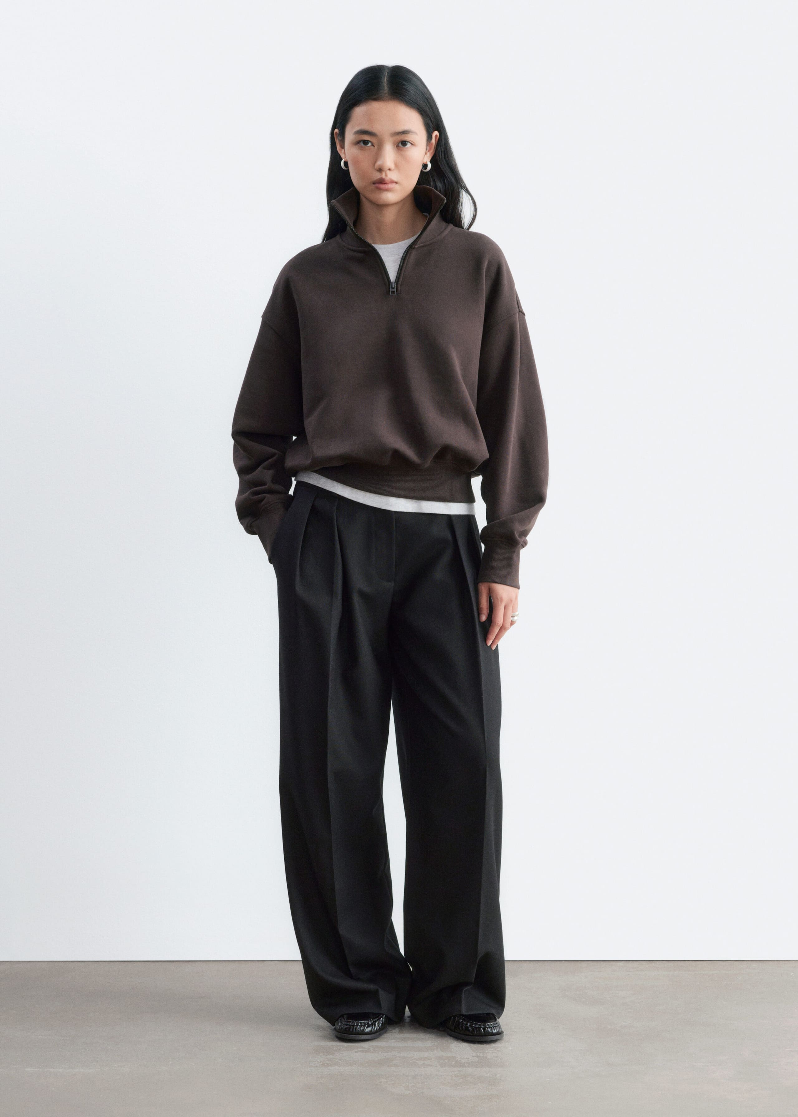 A dark brown half-zip sweatshirt is worn by the model, featuring a high collar, ribbed cuffs and a ribbed hem. The relaxed-fit sweatshirt is layered over a white T-shirt and styled with black wide-leg trousers.