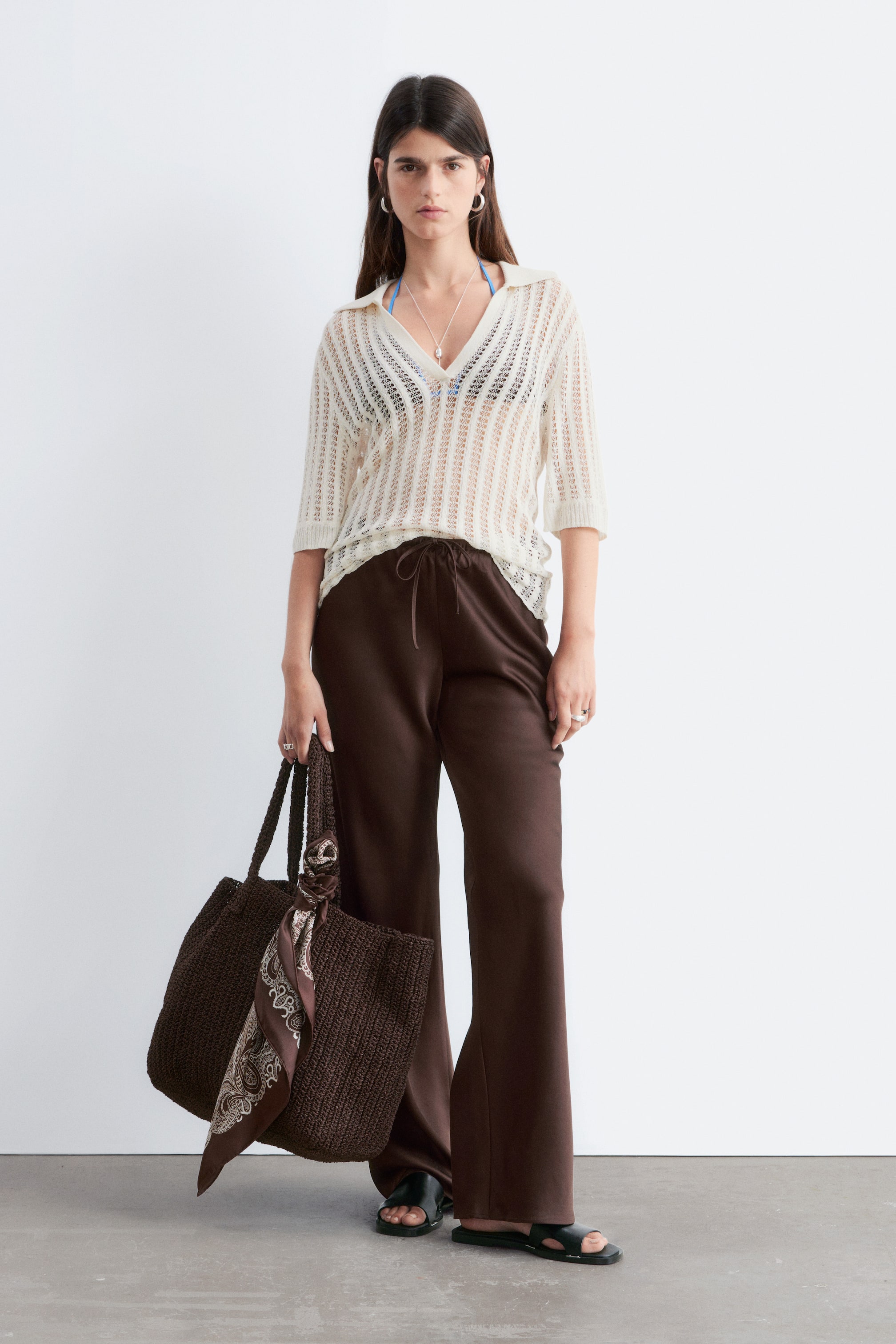 Vis større bilde: En brun vevd tote-veske holdes, med en synlig teksturert vev og to tykke håndtak. Et mønstret silkeskjerf er knyttet rundt ett av håndtakene.