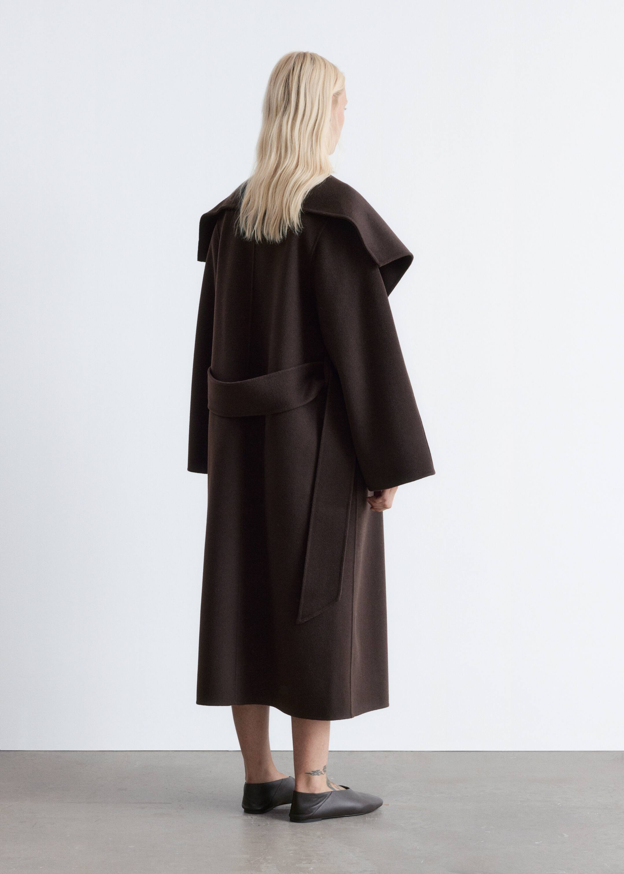 Visualizza immagine più grande: Un cappotto marrone scuro viene visto dal lato posteriore destro, mostrando un grande colletto drappeggiato che si estende sulle spalle. La vestibilità ampia del cappotto è stretta in vita da una cintura dello stesso tessuto annodata sul retro, mentre sono visibili le maniche larghe e lunghe fino al polso e l'orlo raggiunge la lunghezza midi.