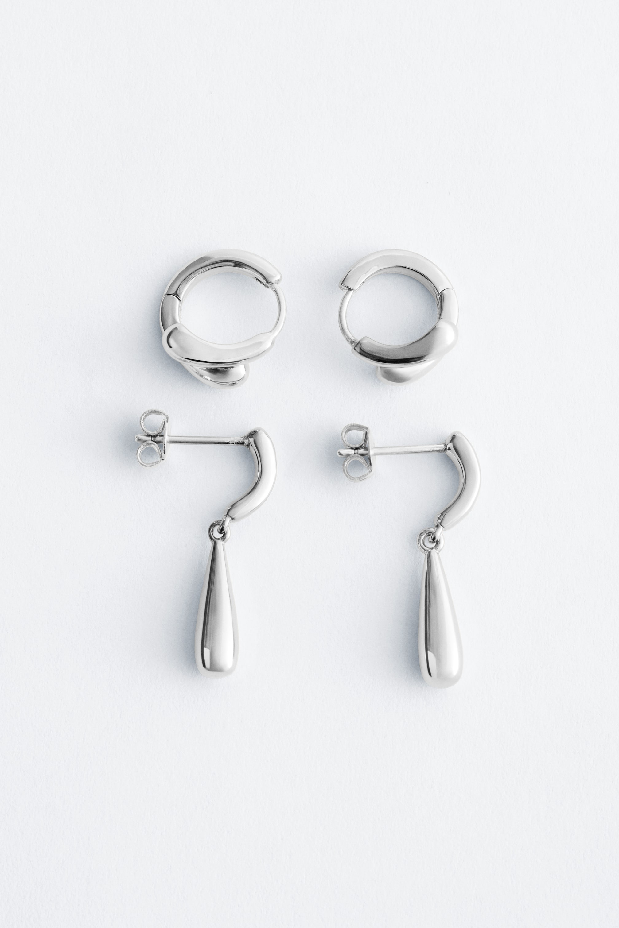 Agrandir l'image: Deux paires de boucles d'oreilles argentées sont exposées. La paire du haut présente des créoles polies avec un nœud de forme organique. La paire du bas montre des clous d'oreilles avec une tige courbée et un pendentif en forme de goutte, avec une tige et un fermoir papillon visibles.