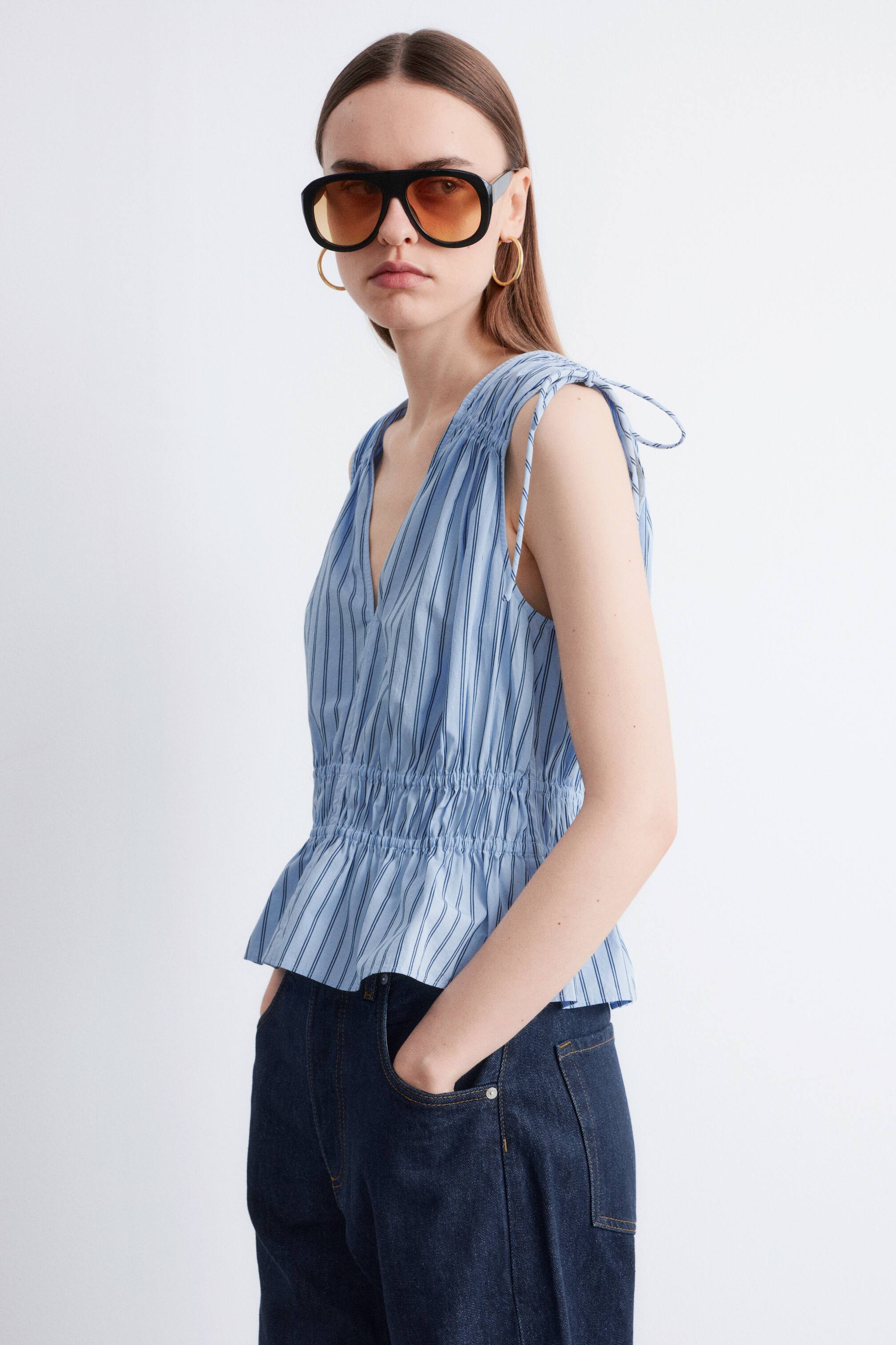 Grotere afbeelding bekijken: Een lichtblauw en wit gestreepte mouwloze top, met een V-hals, elastische tailleplooien en schoudertrekkoorden, wordt gedragen door het model, dat ook een donkere jeans draagt.