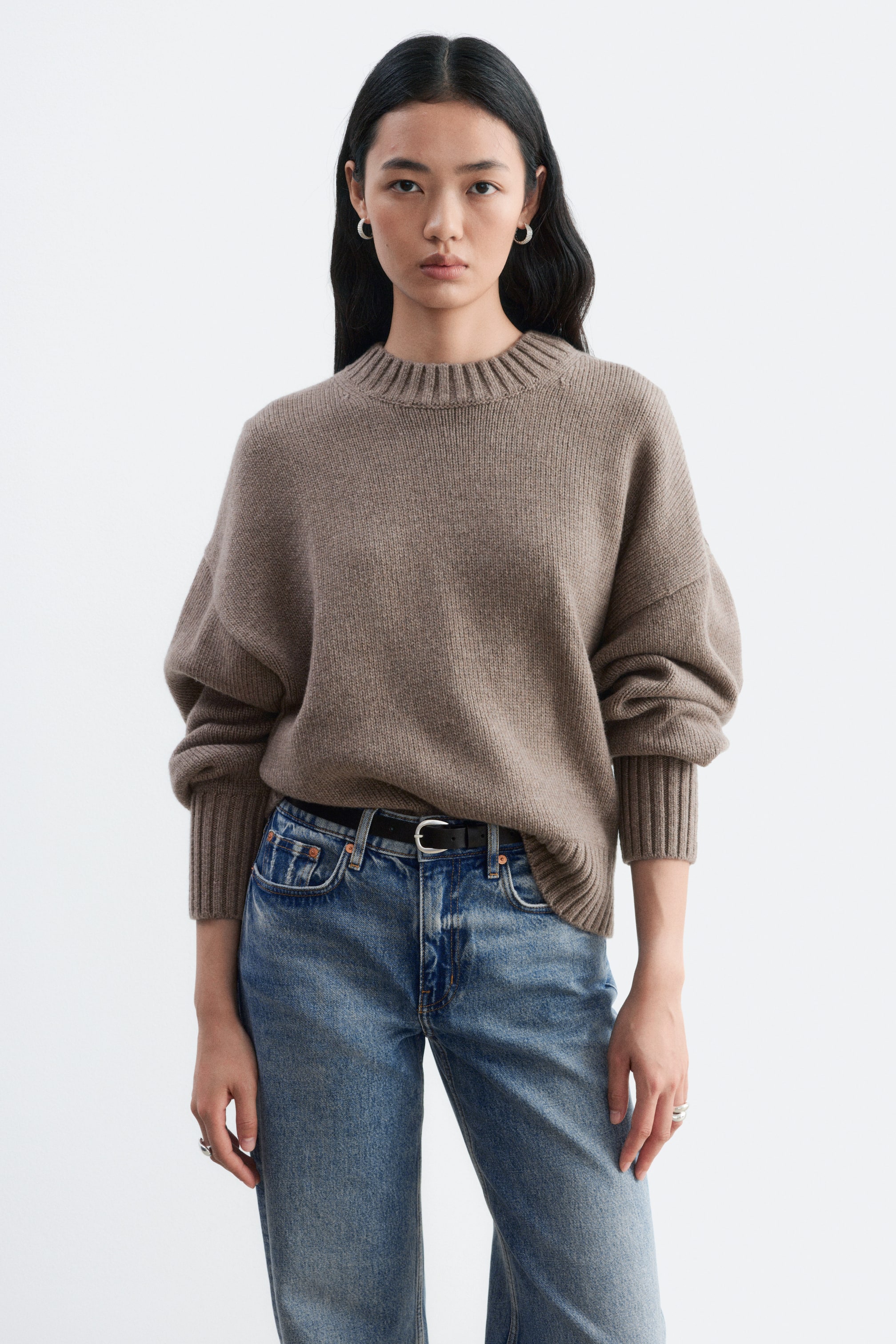 View larger image: A taupe, oversized crew neck jumper is being worn by the model, featuring dropped shoulders and wide ribbed trims on the collar and cuffs. The loose-fitting knitwear is partially tucked at the front into a pair of blue jeans with a black belt.