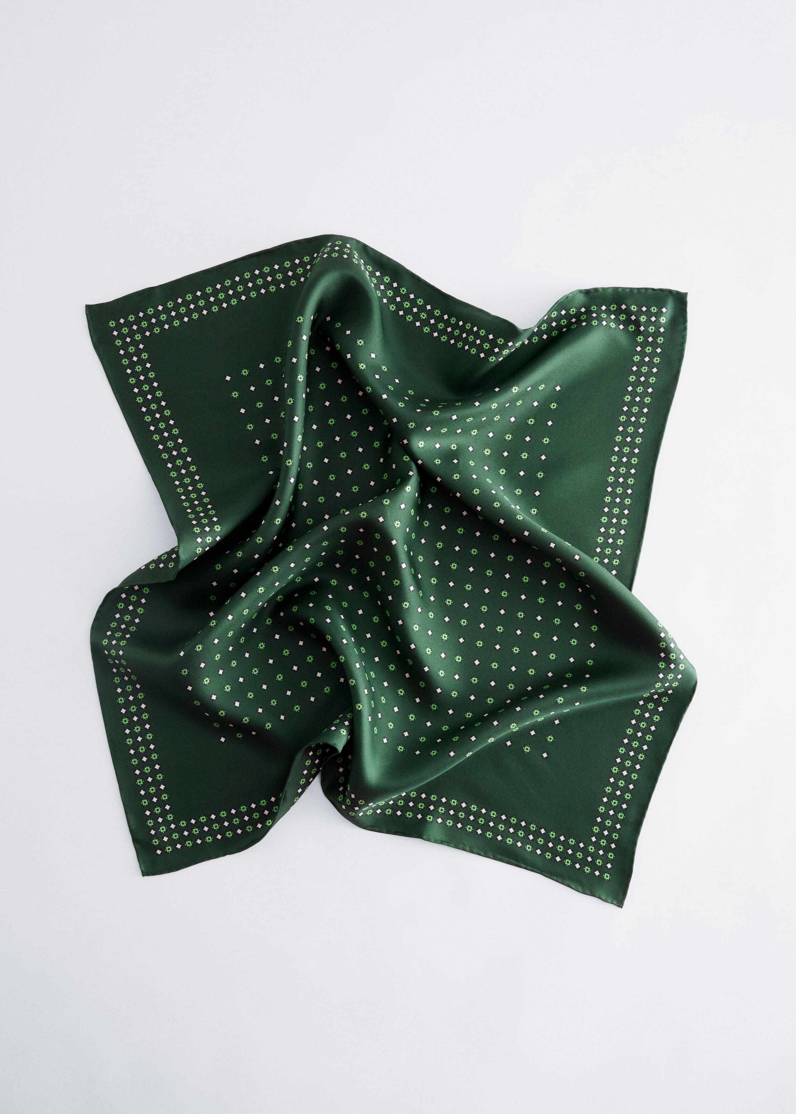 A dark green square scarf, made of lustrous, smooth fabric, is styled with soft folds to highlight its sheen. It features a repeating pattern of small pink and white geometric diamonds across the center and along its double-row border.