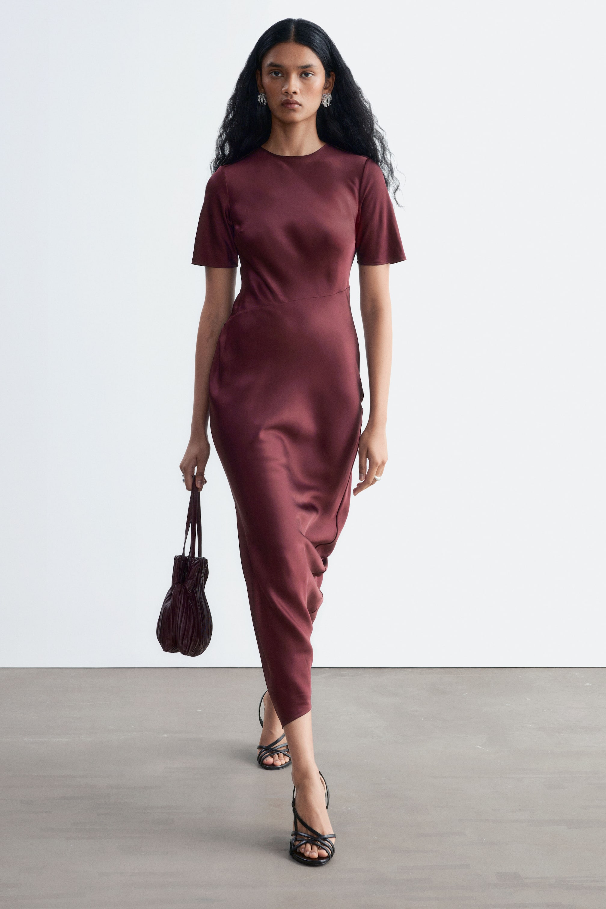 Visualizza immagine più grande: La modella indossa un abito in raso color bordeaux, lungo fino alla caviglia, con scollo rotondo e maniche corte. L’abito slim fit è abbinato a sandali neri con tacco e cinturini e a una borsa coordinata con dettaglio arricciato, tenuta in una mano.