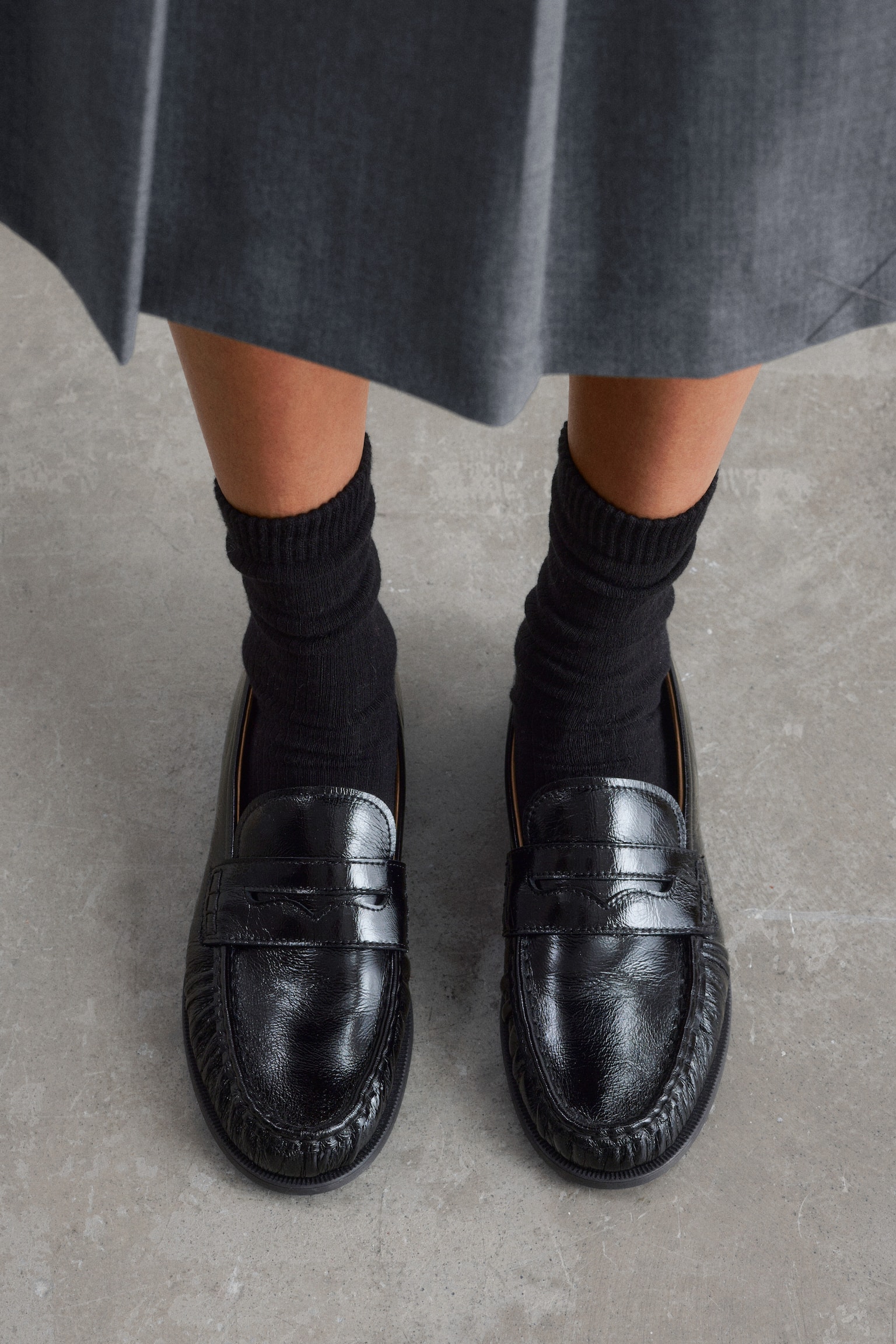 En svart, høyglanset loafer blir presentert fra en sidevisning, vinklet litt mot fronten. Den har en firkantet moc-tå med fremtredende sømmer og en klassisk penny loafer-reim over vristen, og støttes av en lav, flat hæl. - 1