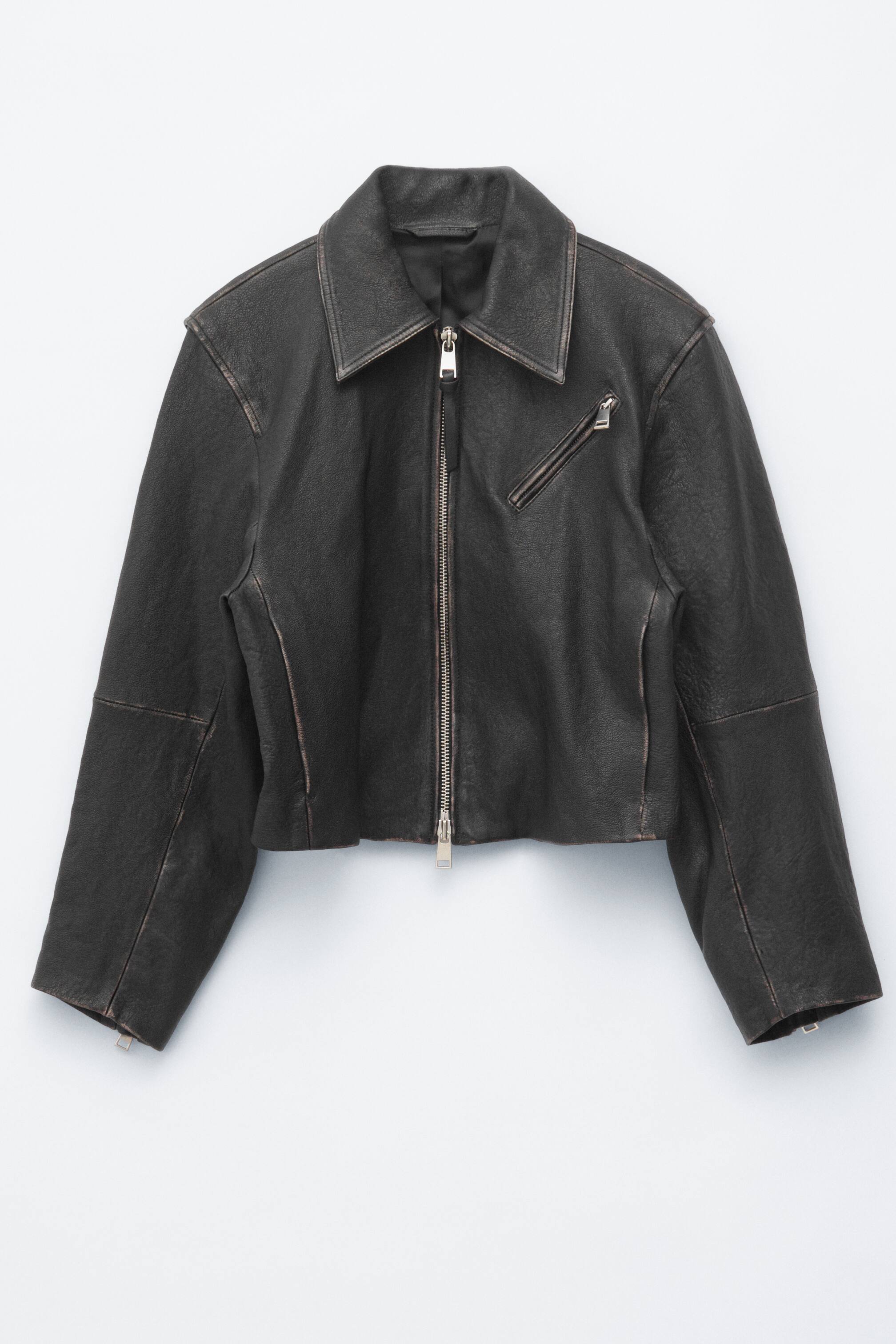 Grotere afbeelding bekijken: Een kort, boxy jasje in donkerbruin mat leer met een gedragen uitstraling, voorzien van gestructureerde schouders, een klassieke kraag en een centrale zilverkleurige ritssluiting. Het is gedetailleerd met een schuin geplaatste zilverkleurige ritszak op de linkerborst, snijlijnnaden en ritssluitingen aan de mouwen.