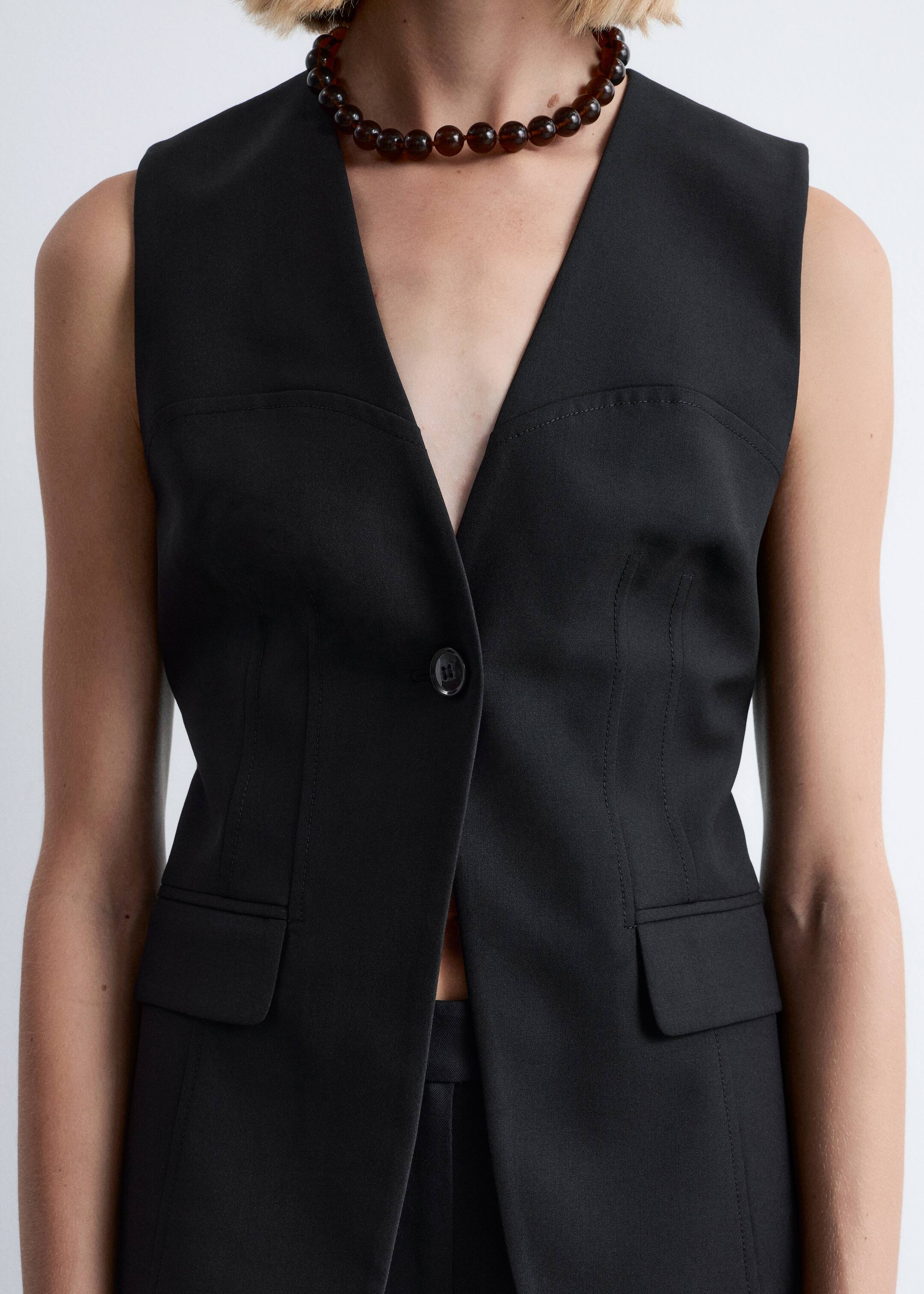 Agrandir l'image: Un gilet sans manches noir est montré de face. Il présente un décolleté en V profond et une fermeture à un seul bouton, actuellement déboutonnée. Le gilet est conçu avec deux poches à rabat aux hanches et des coutures de façonnage sur mesure, y compris des coutures horizontales au niveau de la poitrine et des coutures verticales princesse, pour une coupe structurée.