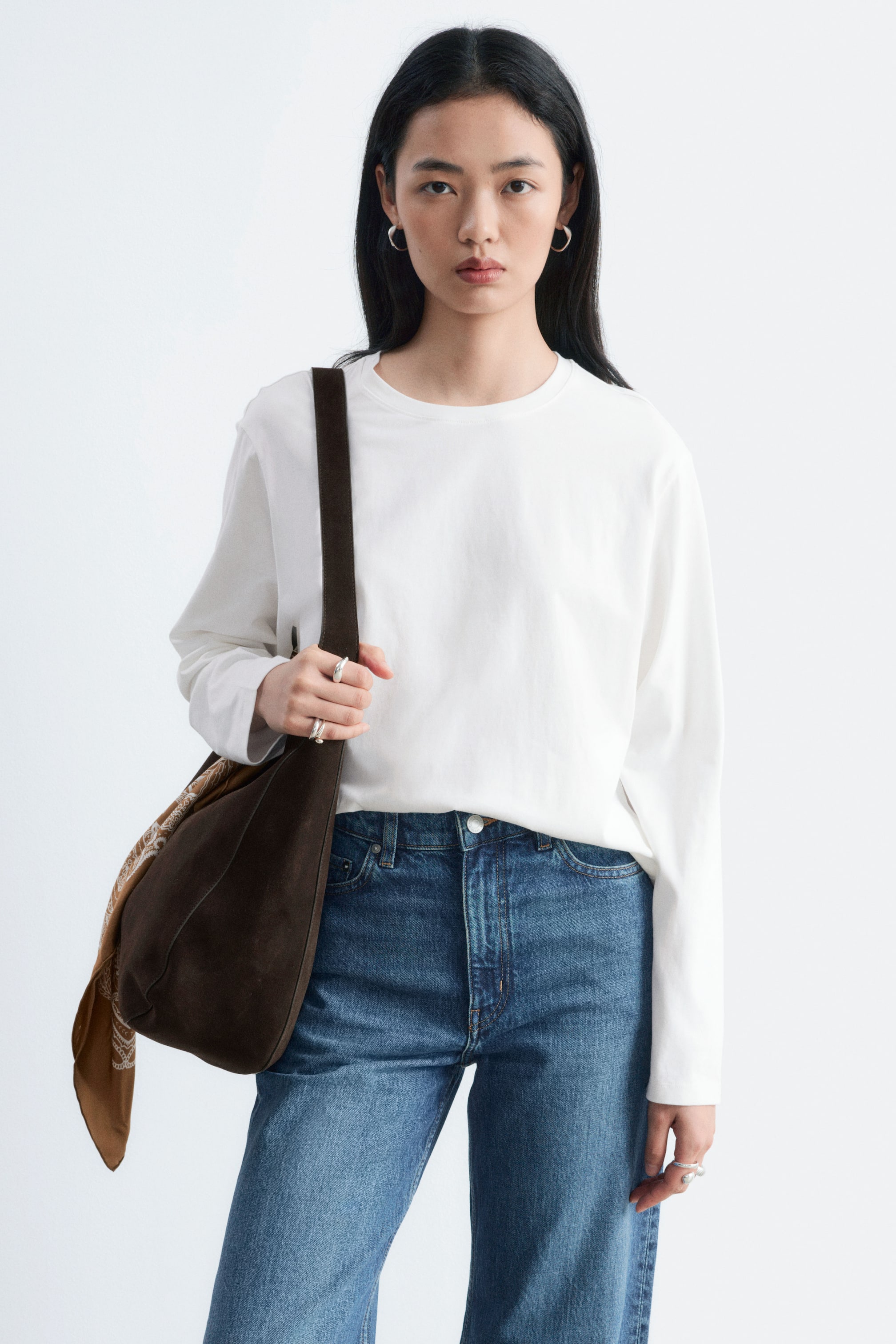 Visualizza immagine più grande: La modella indossa un top bianco a maniche lunghe con scollo tondo, infilato in un paio di jeans blu. Il top dalla vestibilità rilassata è abbinato a una borsa a spalla in suede marrone scuro, portata a tracolla.