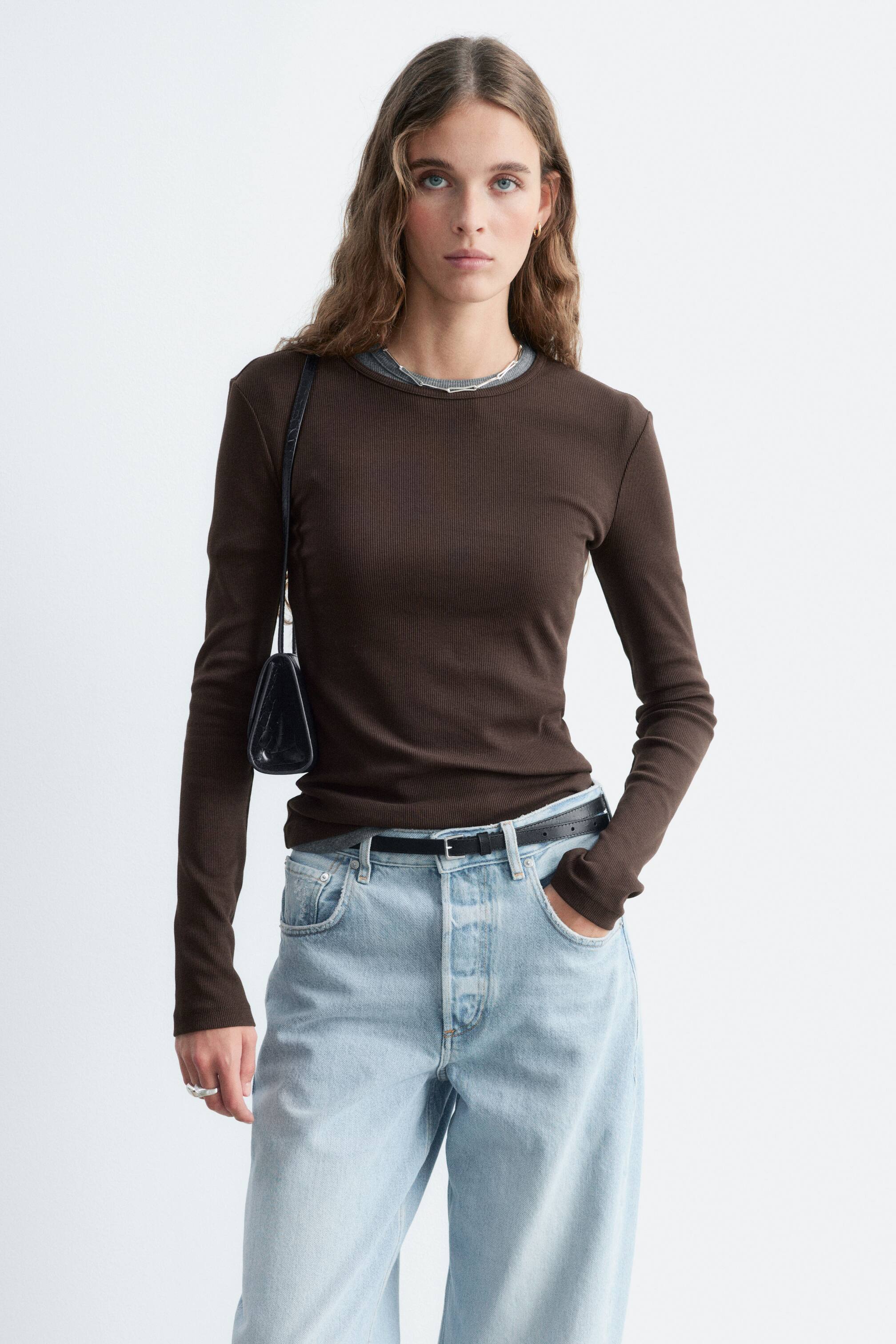 Visa större bild: En mörkbrun, ribbad långärmad topp bärs av modellen, lager på lager över en grå rundhalsad krage och instoppad i ljusblå jeans. Den slim fit-passande toppen är stylad med ett tunt svart bälte och en svart axelväska.