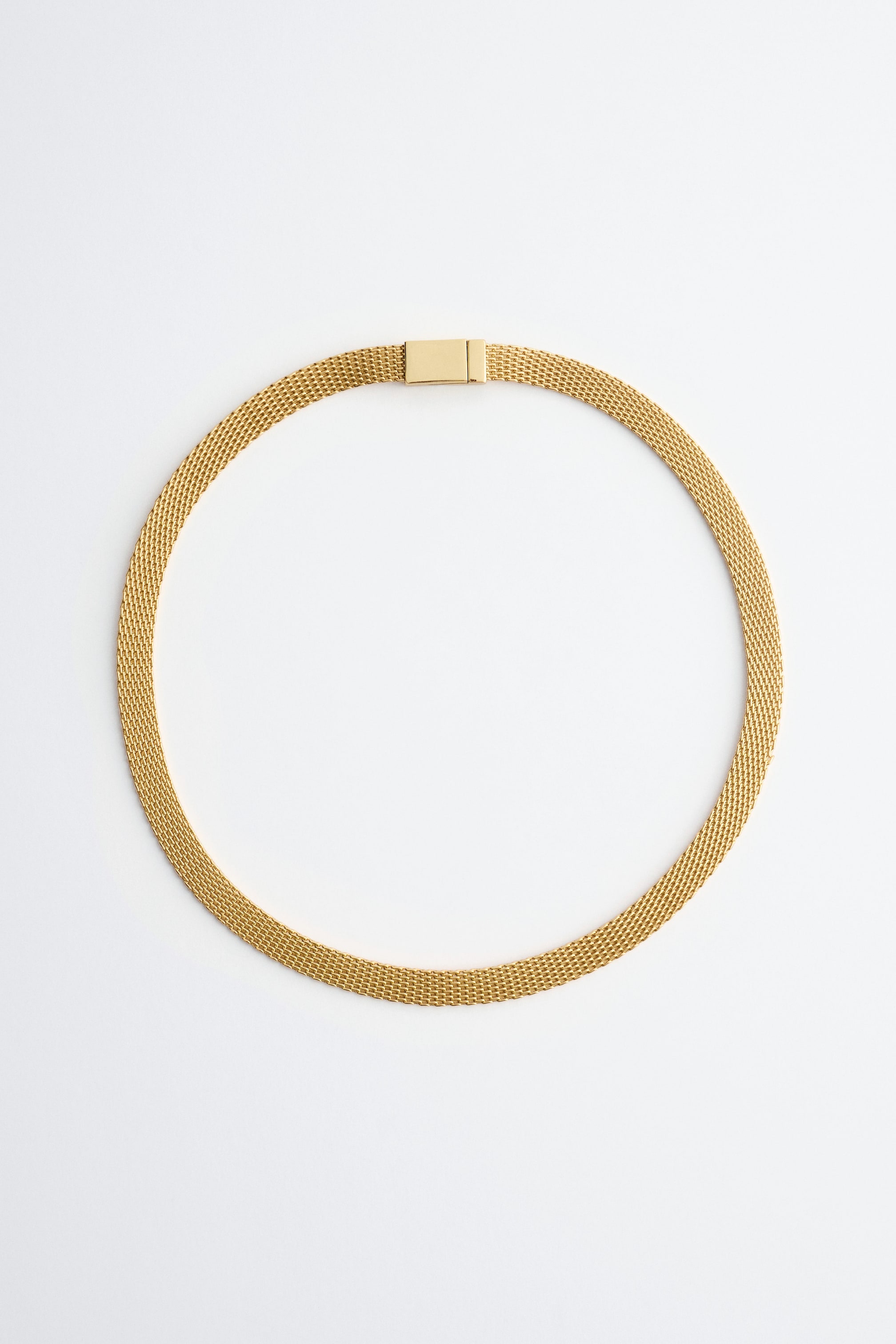 View larger image: A polished brass flat chain necklace features a mesh-like texture and a rectangular clasp, presented in a circular shape.