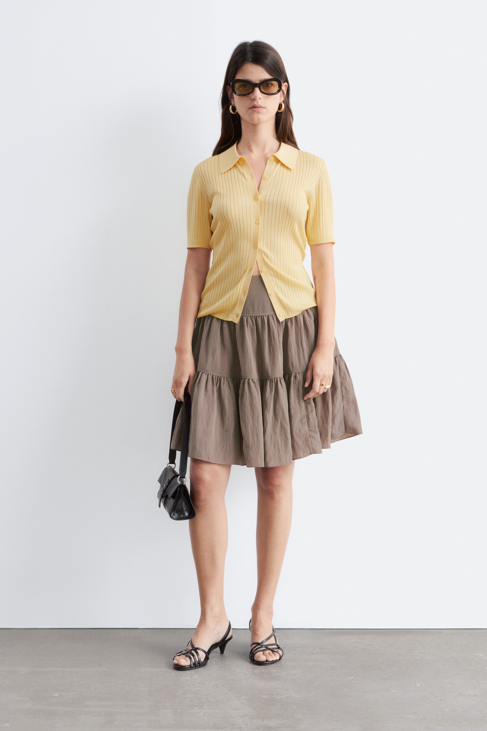 Agrandir l'image: La modèle porte une jupe trapèze taupe à volants, longueur genou. Elle présente des couches douces créant du volume et est assortie d'un haut jaune à manches courtes et de talons noirs à brides.