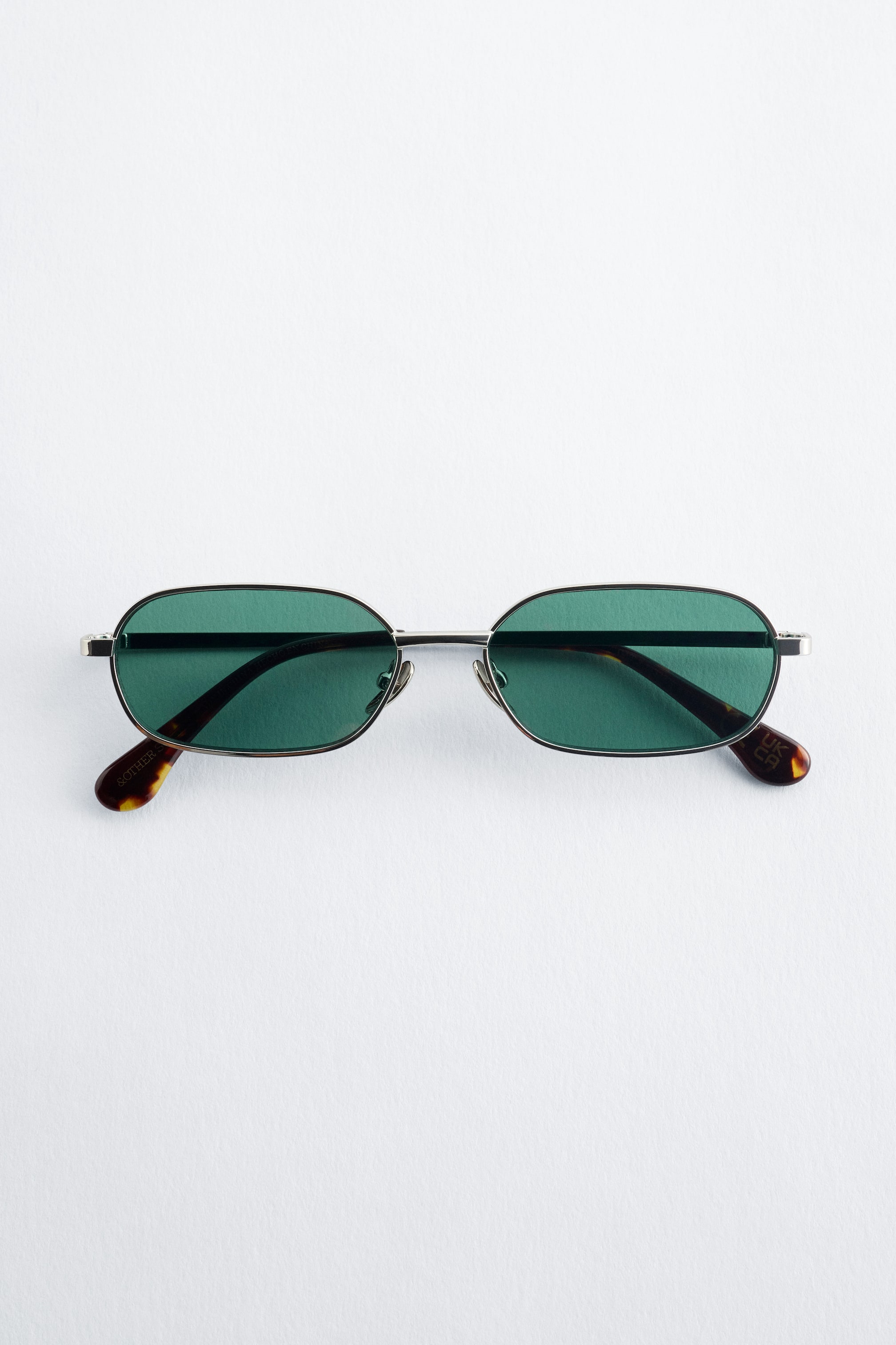 Ver imagen más grande: Estas gafas de sol presentan una montura delgada de acero inoxidable en tono plateado. La montura sostiene lentes pequeñas y rectangulares de color verde con bordes suavemente redondeados, y las patillas muestran puntas de concha de tortuga en tono marrón oscuro.