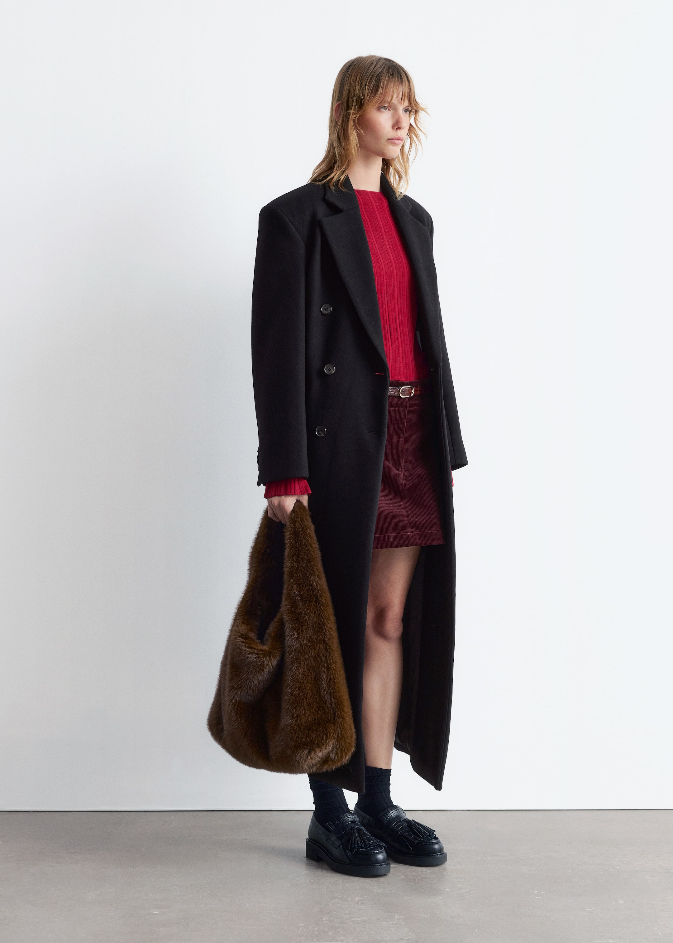 View larger image: A long black double-breasted coat with notch lapels is being worn open in a three-quarter view, revealing a red ribbed long-sleeve top and a burgundy corduroy mini-skirt. The ensemble is completed with black chunky loafers, dark socks, and a large brown faux fur bag being held.
