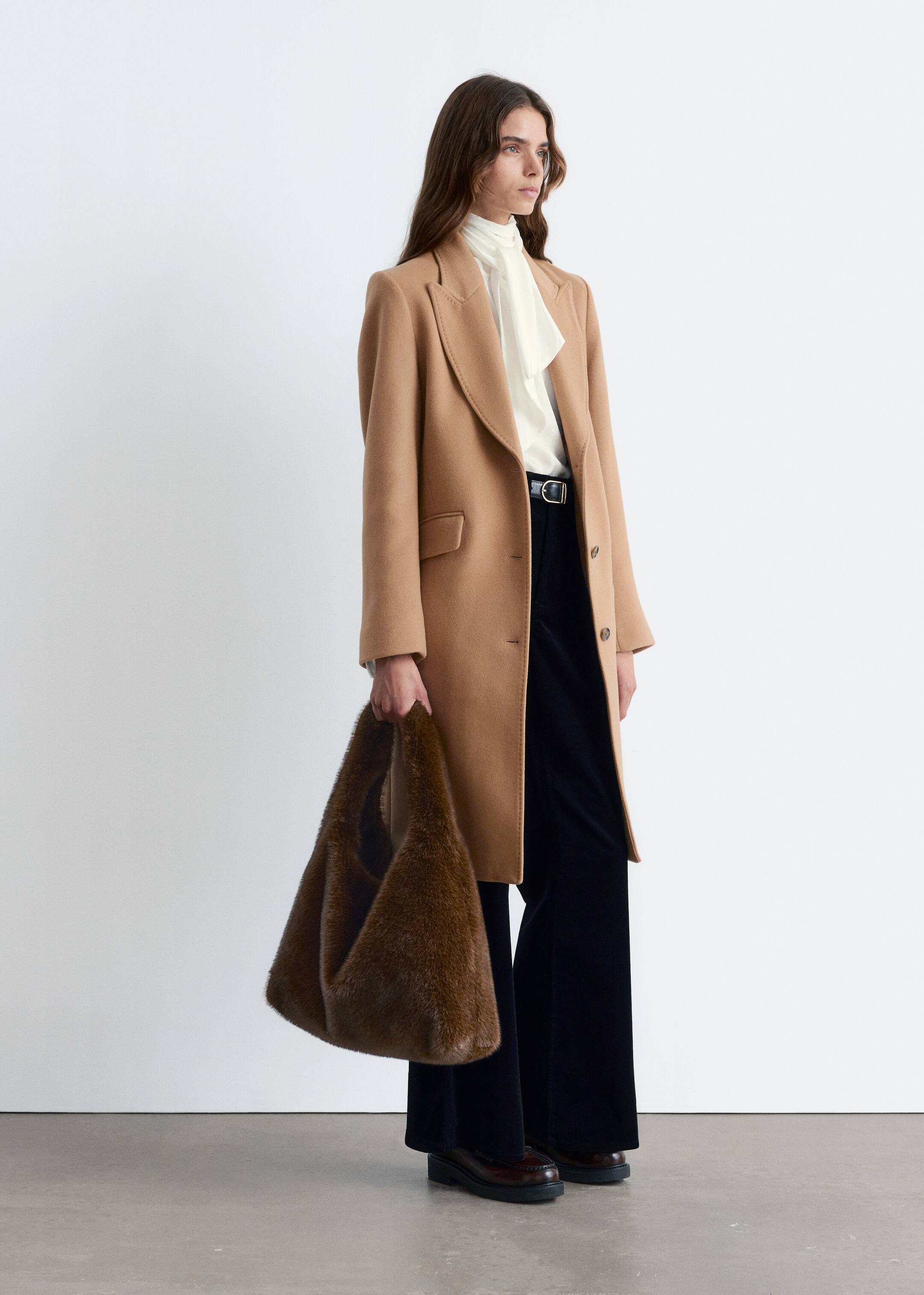 View larger image: A camel-colored single-breasted coat is being shown from a slight side angle, displaying notch lapels, three visible buttons, and flap pockets. It is cut with a straight fit and falling to approximately mid-thigh length.