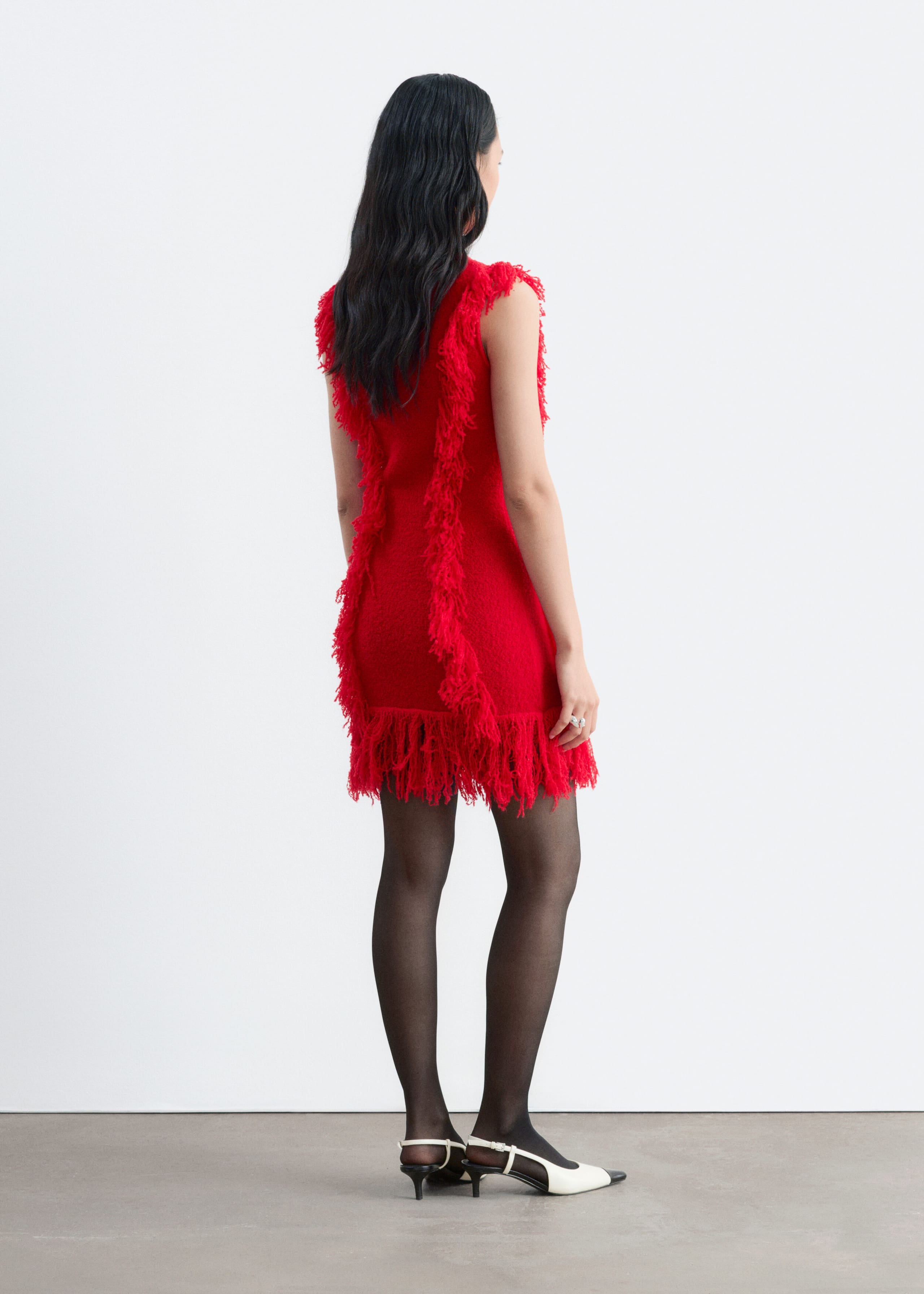 A red, sleeveless, slim-fit dress featuring vertical rows of textured fringe and a fringed hem is being worn by the model. The dress is styled with sheer black tights and black-and-white slingback heels.
