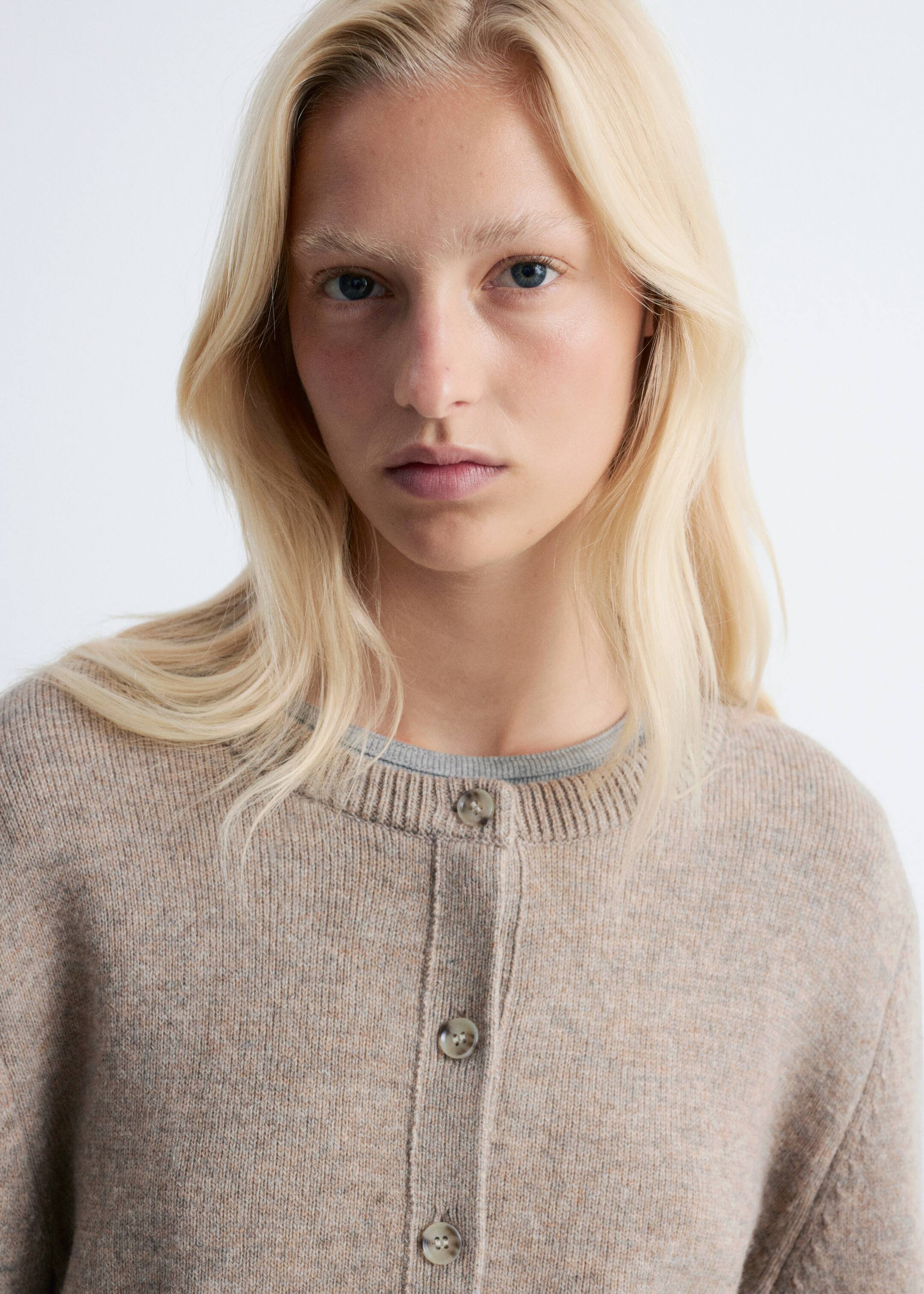 View larger image: A heathered beige knitted cardigan is being worn, featuring a ribbed round neckline and a button placket with three light-colored, round buttons. A grey crewneck t-shirt is visible underneath the cardigan's neckline.