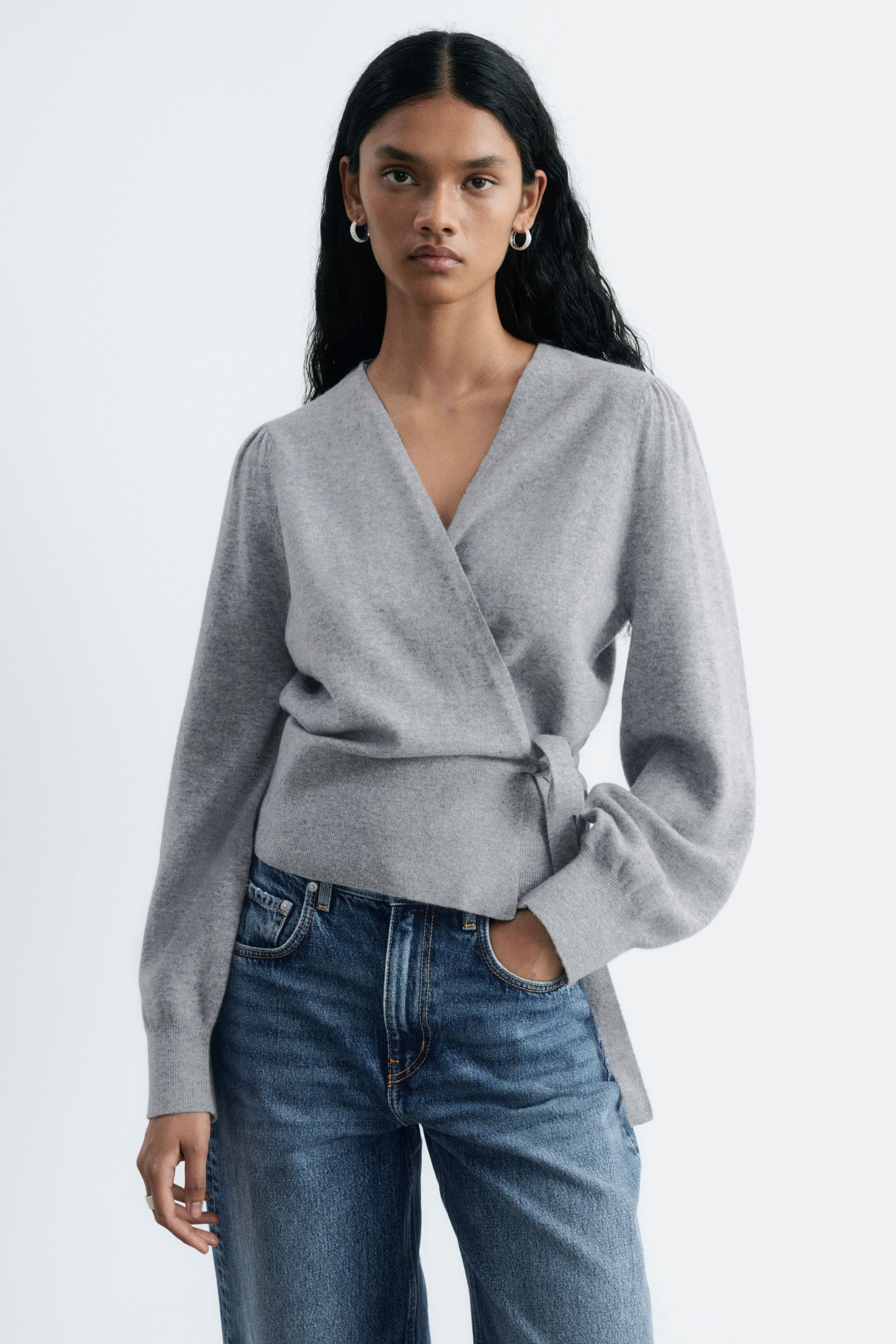 Visualizza immagine più grande: La modella indossa un cardigan incrociato grigio. Presenta scollo a V, dettaglio con cintura annodabile in vita, maniche lunghe a palloncino e polsini e fondo a costine. La modella sta infilando una mano nella tasca dei jeans.