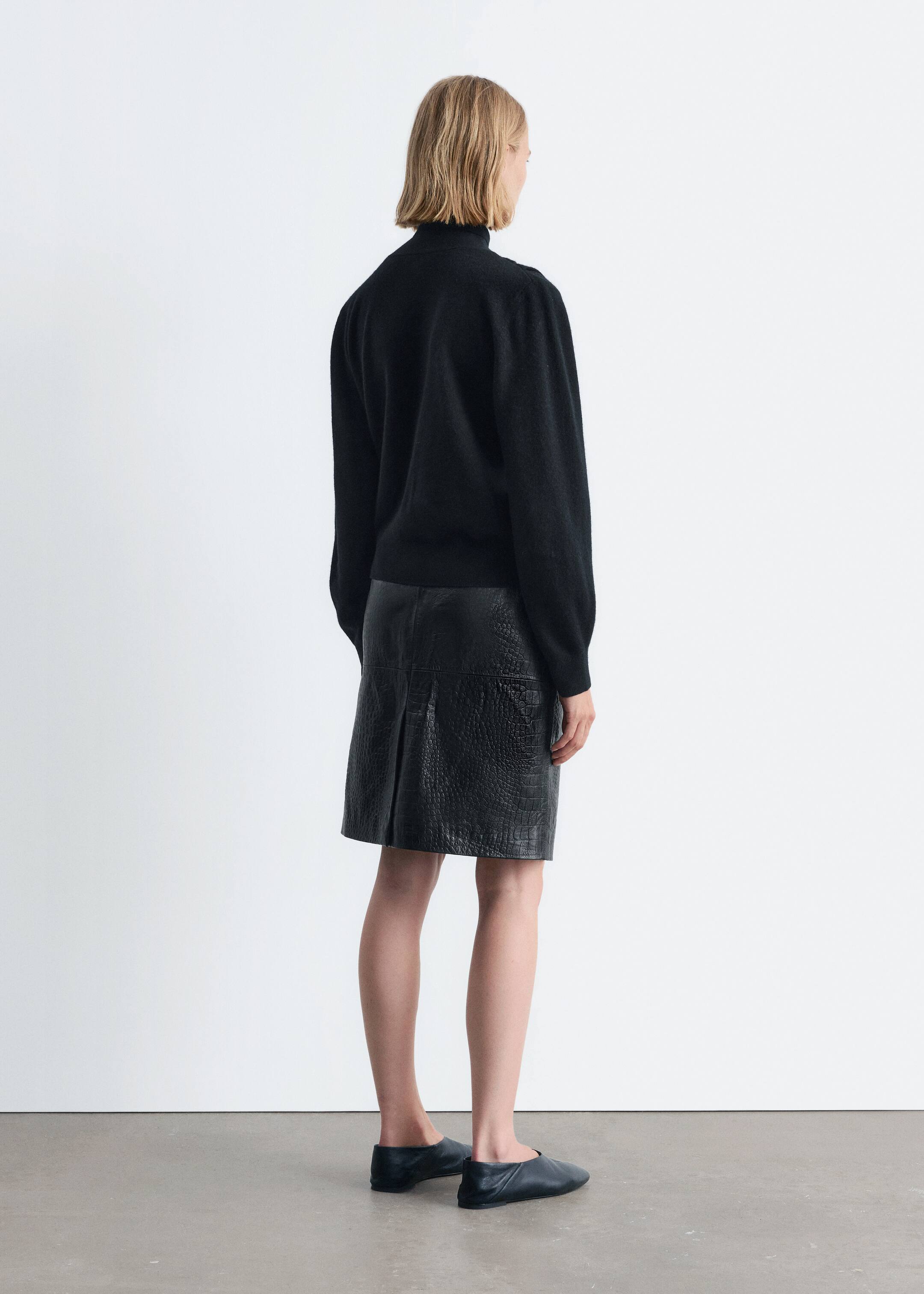 Größeres Bild ansehen: Ein schwarzer Rollkragenpullover mit subtiler Schultervolumen und einem eng anliegenden Saum wird getragen und von hinten betrachtet. Er wird mit einem schwarzen knielangen Rock mit geprägter Krokodilstruktur und einer zentralen Rückfalte gestylt, ergänzt durch schwarze flache Lederschuhe.