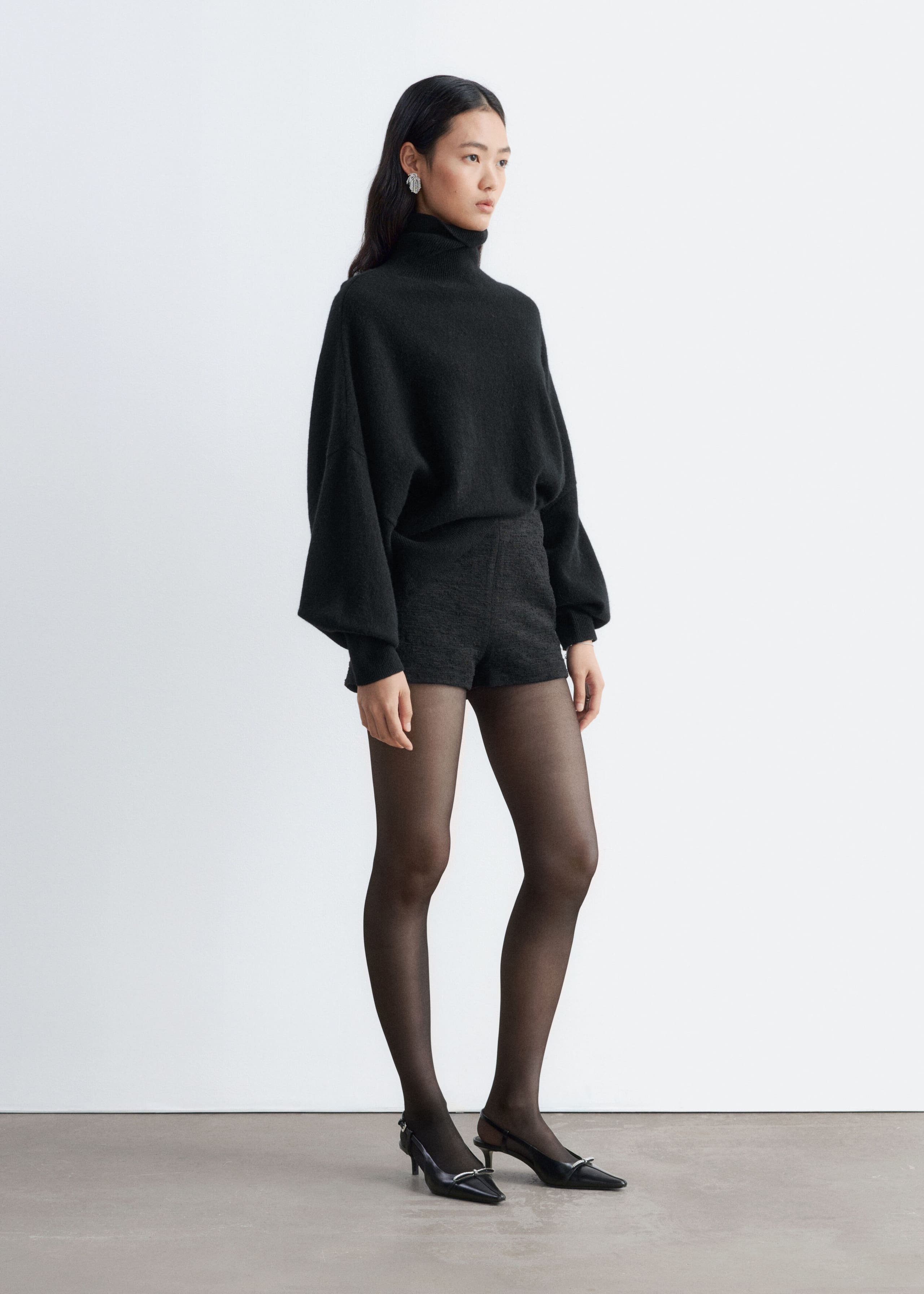 These dark grey tweed shorts are modelled on a high waist, structured silhouette and textured fabric. They are styled with a tucked-in black roll-neck sweater, sheer black tights, and black pointed-toe slingback heels.
