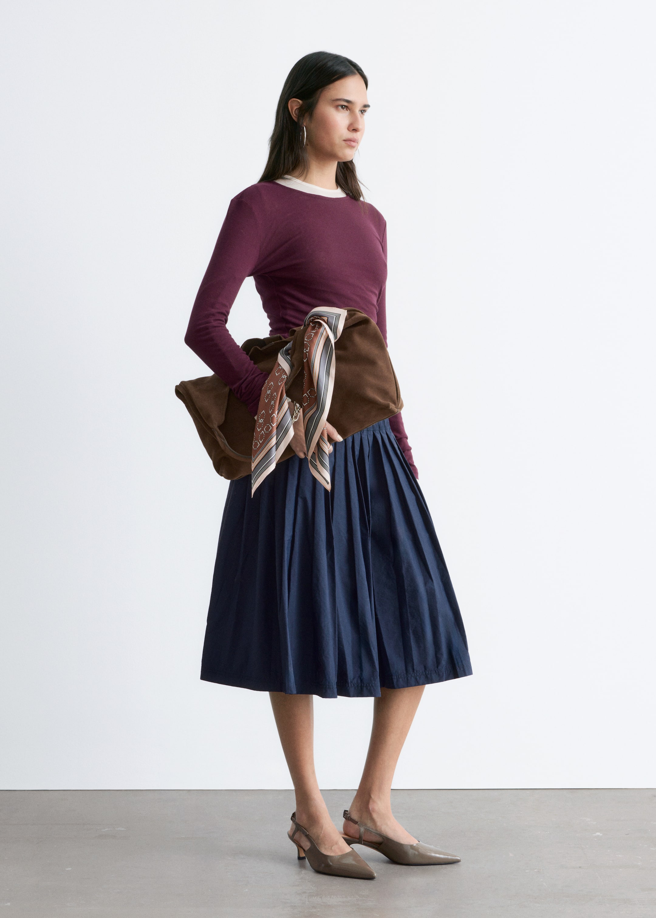 Größeres Bild ansehen: Ein bordeauxrotes Langarm-Top mit einer cremefarbenen Rundhalsausschnitt-Umrandung wird präsentiert, in einen marineblauen plissierten Midirock gesteckt. Eine braune, weich strukturierte Schultertasche mit einem gemusterten Schal, der an den Riemen gebunden ist, wird an der Hüfte getragen, dazu taupefarbene Slingback-Pumps mit spitzer Zehenpartie.