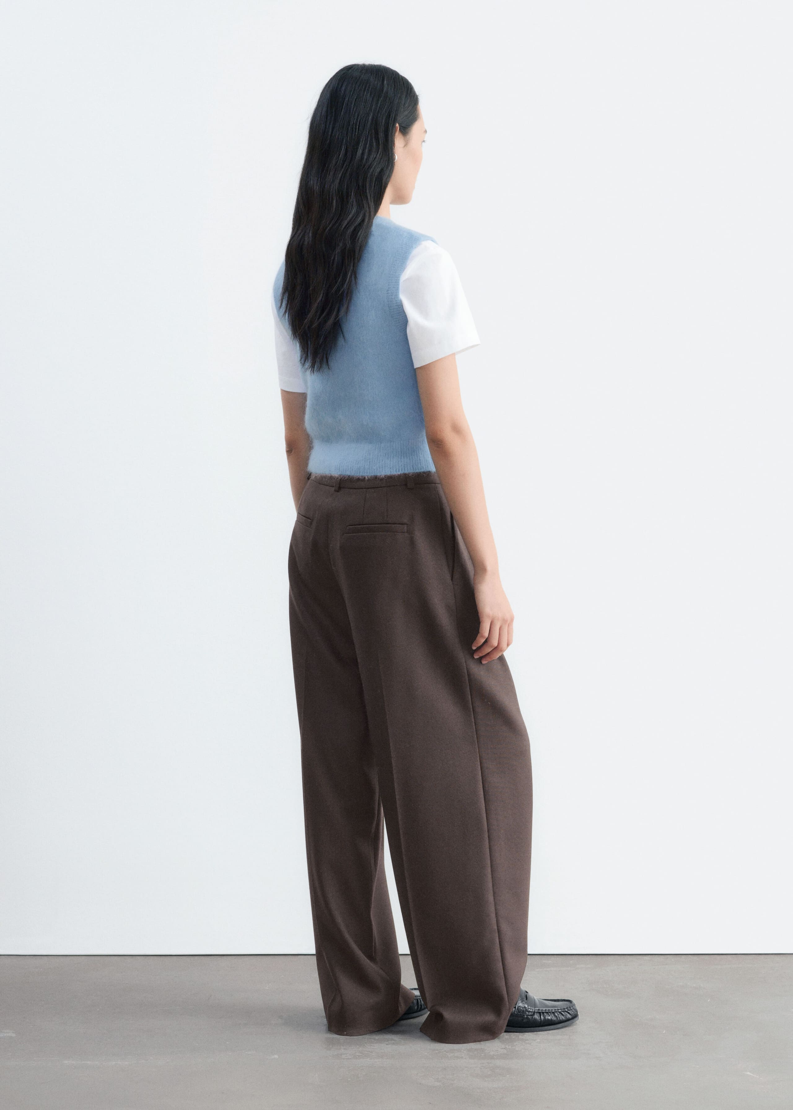 La modelo lleva un chaleco de mezcla de mohair de color azul claro y punto, mostrando su suave superficie texturizada desde un ángulo trasero de tres cuartos. El chaleco se lleva sobre una camiseta blanca de manga corta y se combina con un pantalón marrón de corte holgado.
