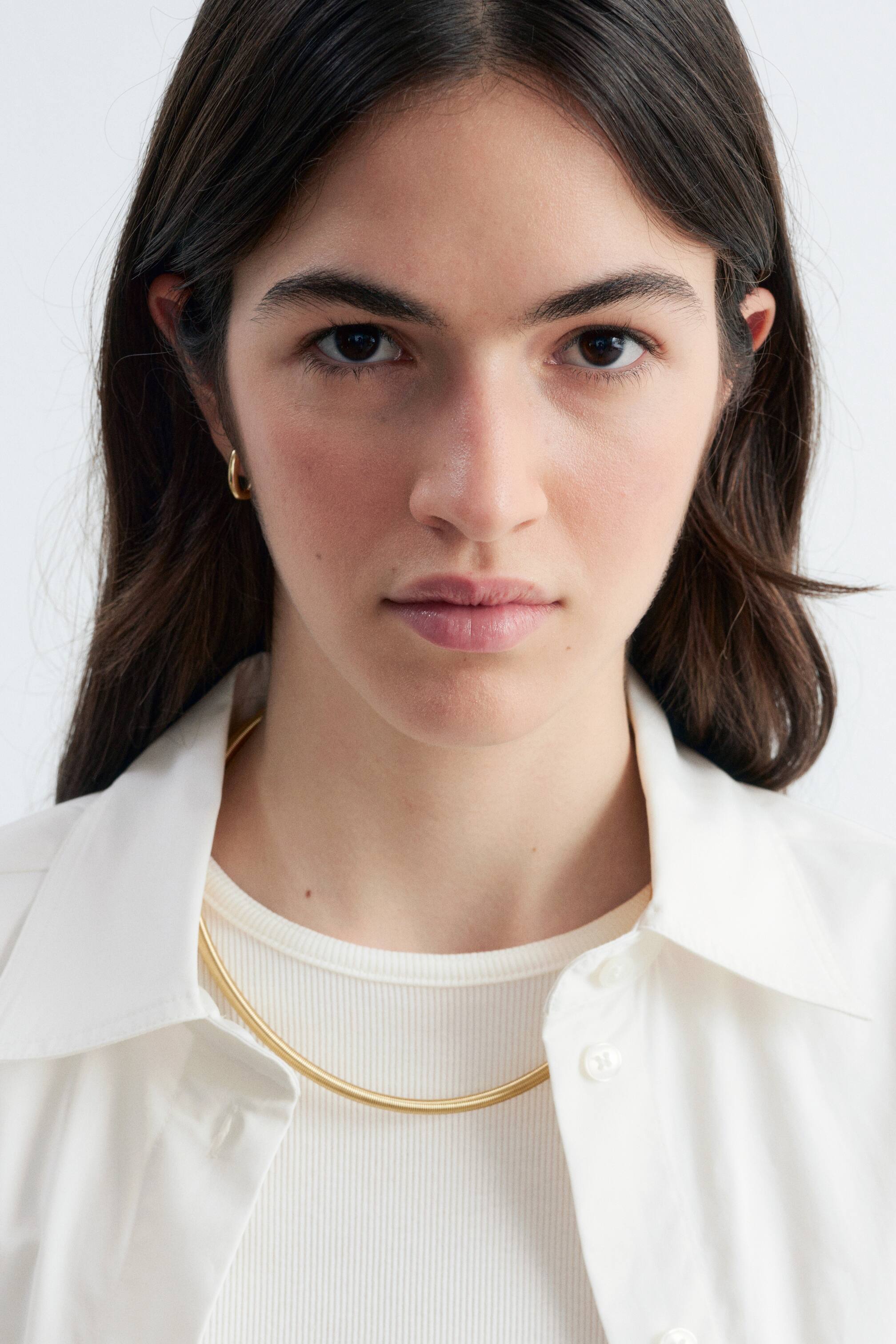 View larger image: A gold-toned snake chain necklace is being worn by the model, resting flat against the neckline of a white ribbed top. The sleek, rounded chain is framed by the open collar of a white shirt.