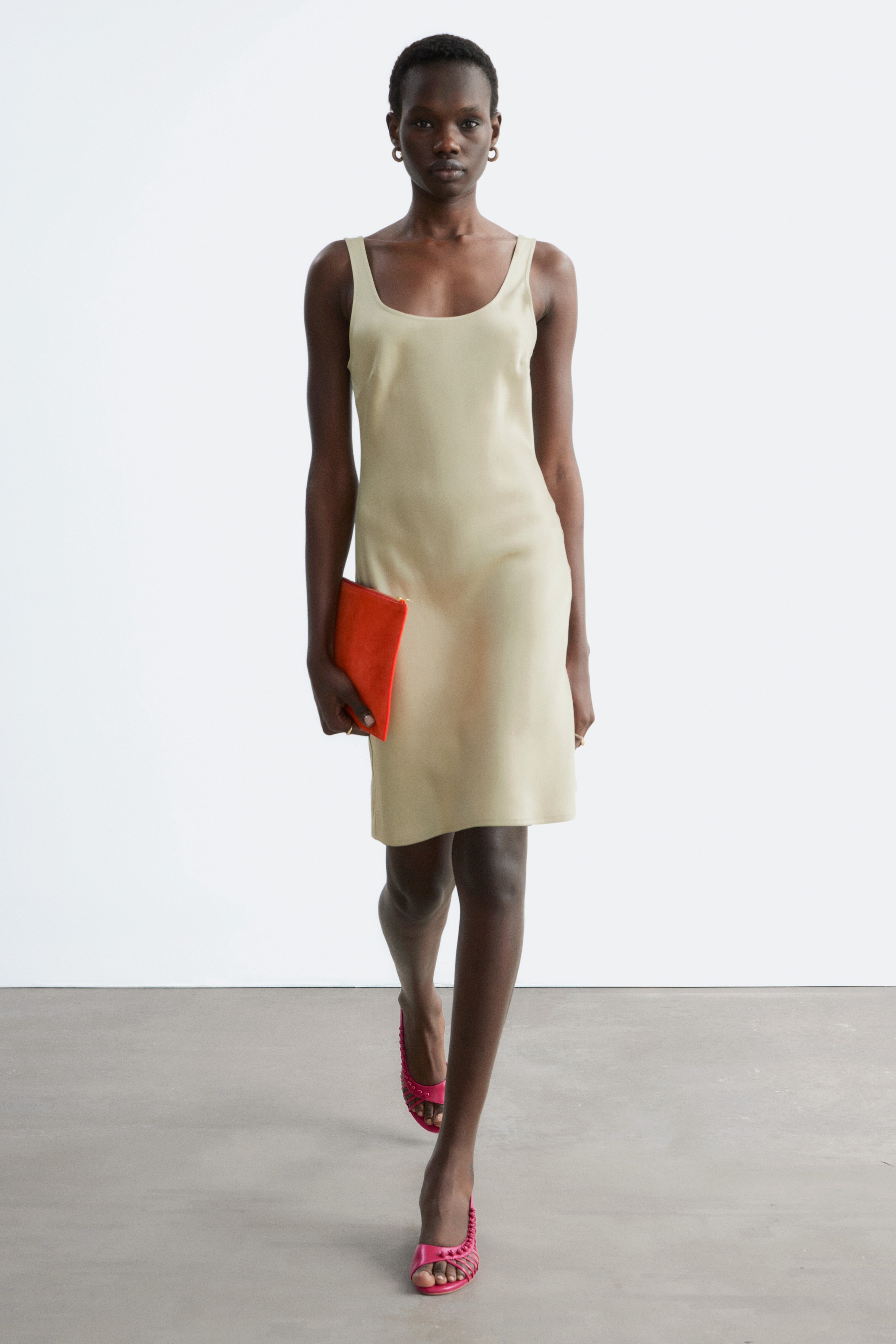 Преглед на по-голямо изображение: Лека бежова блестяща сатенена мини рокля с широки презрамки и овално деколте и гръб, носена от модела. Моделът държи оранжев клъч в едната ръка и носи розови обувки с отворени пръсти.