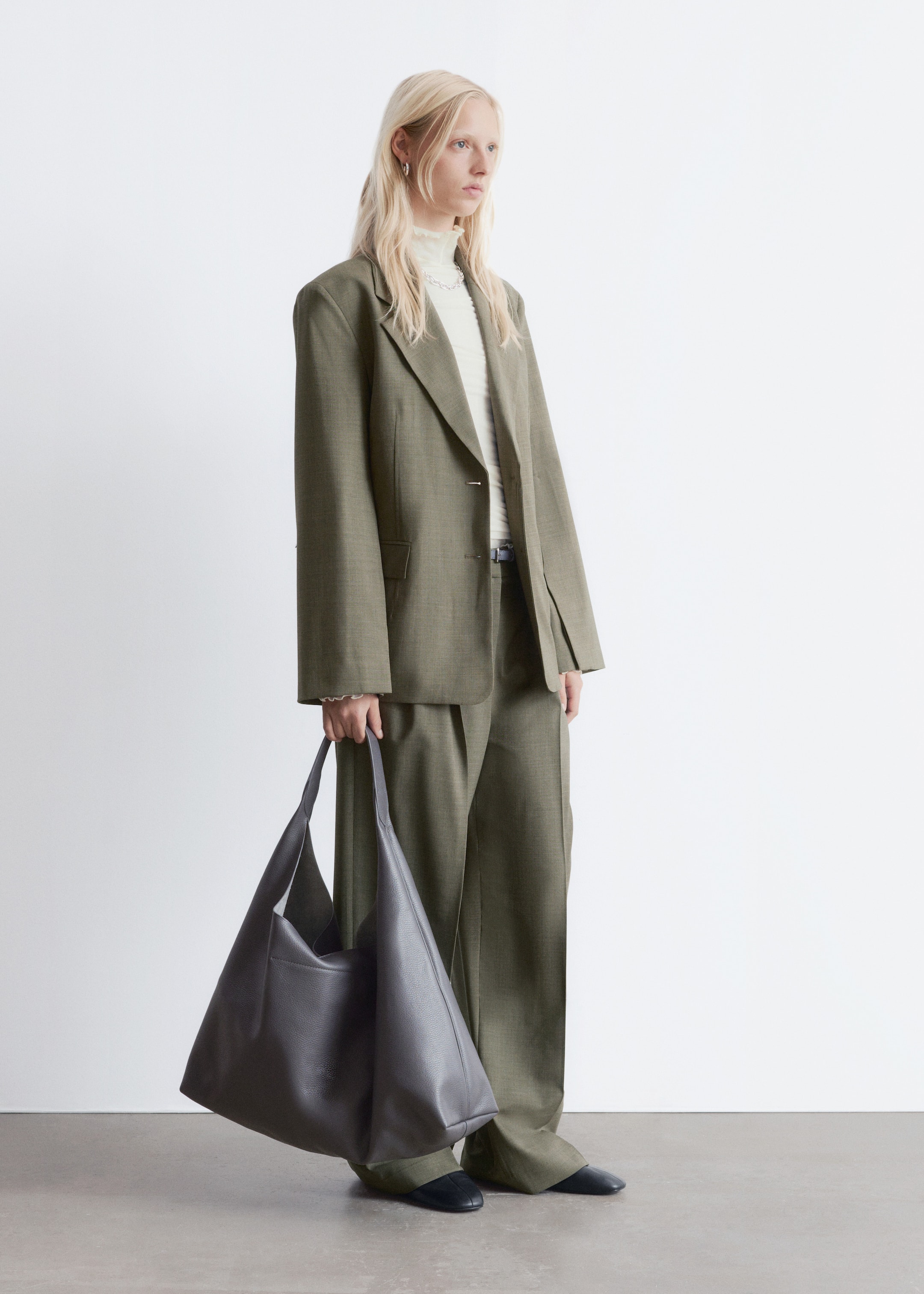 Ingrandisci l'immagine: Una borsa tote in pelle grigia scura, martellata, è tenuta dal suo ampio cinturino, appesa verso il basso. La borsa non strutturata sta formando una forma morbida e leggermente trapezoidale, con la parte anteriore inclinata leggermente verso sinistra.