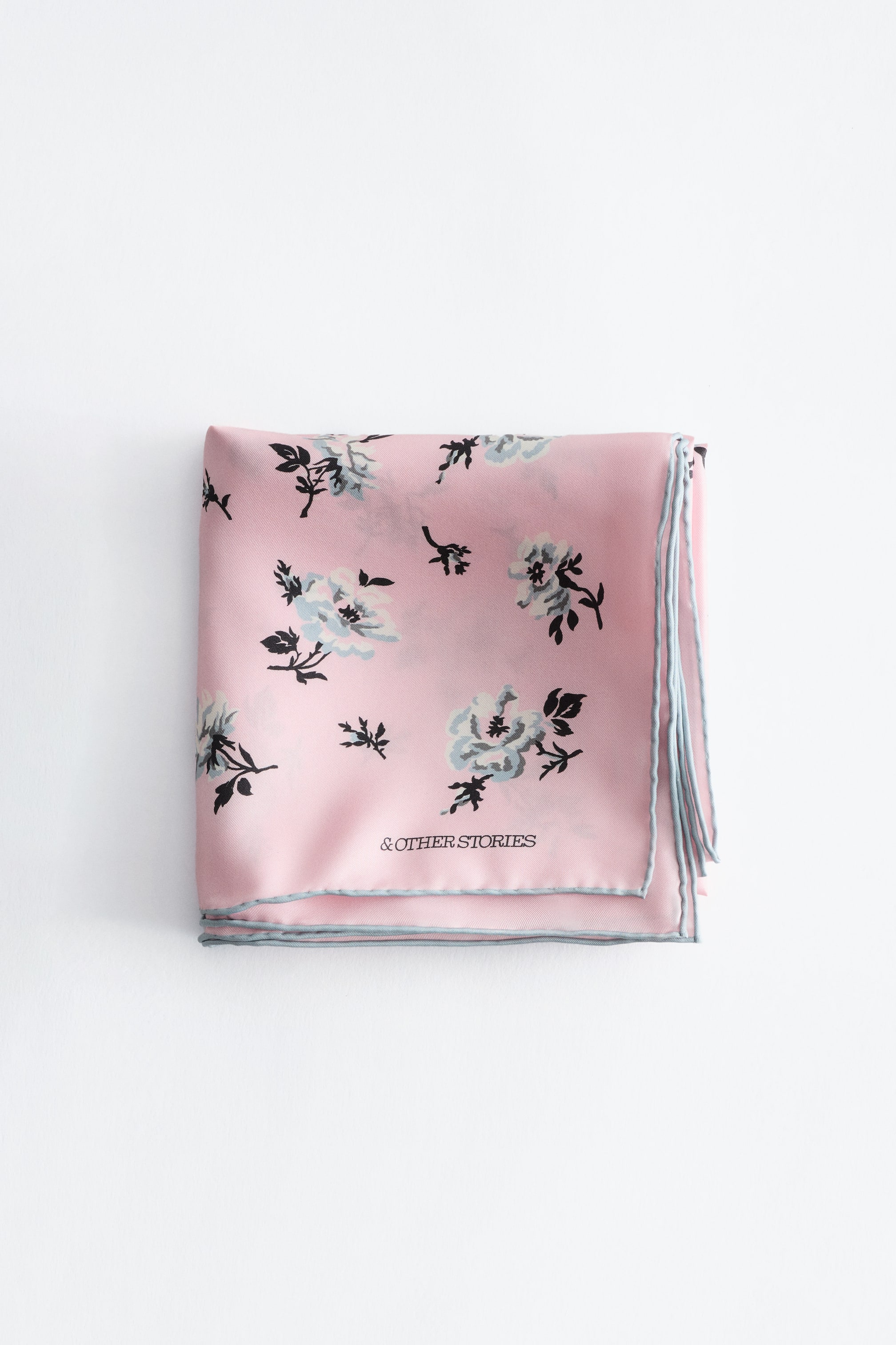 Ver imagem maior: Um lenço de bolso floral rosa está cuidadosamente dobrado, exibindo um padrão de flores e folhas em azul claro e preto. As suas bordas são acabadas com um viés azul claro, e o nome da marca "& OTHER STORIES" está visível na frente.